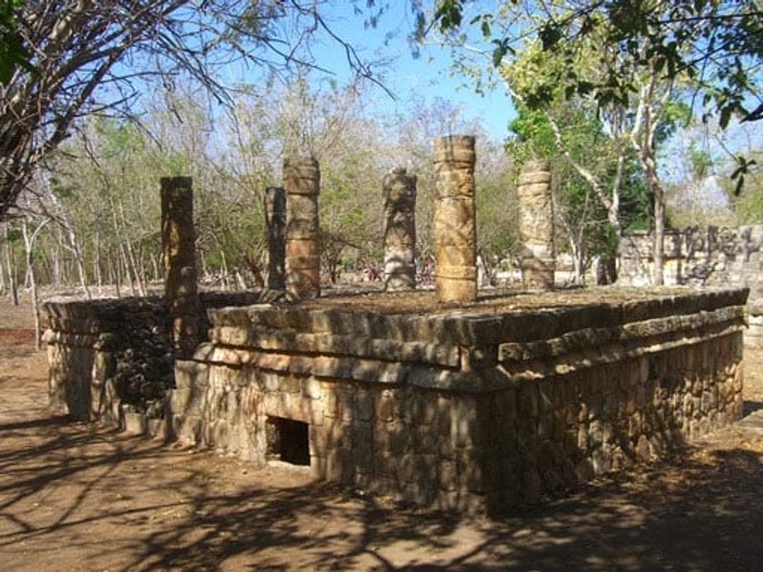 Una antigua estructura de piedra con pilares en la parte superior, ubicada en una zona boscosa. La estructura está bañada por la luz del sol, y los árboles circundantes crean un telón de fondo natural, proyectando sombras moteadas sobre el suelo.