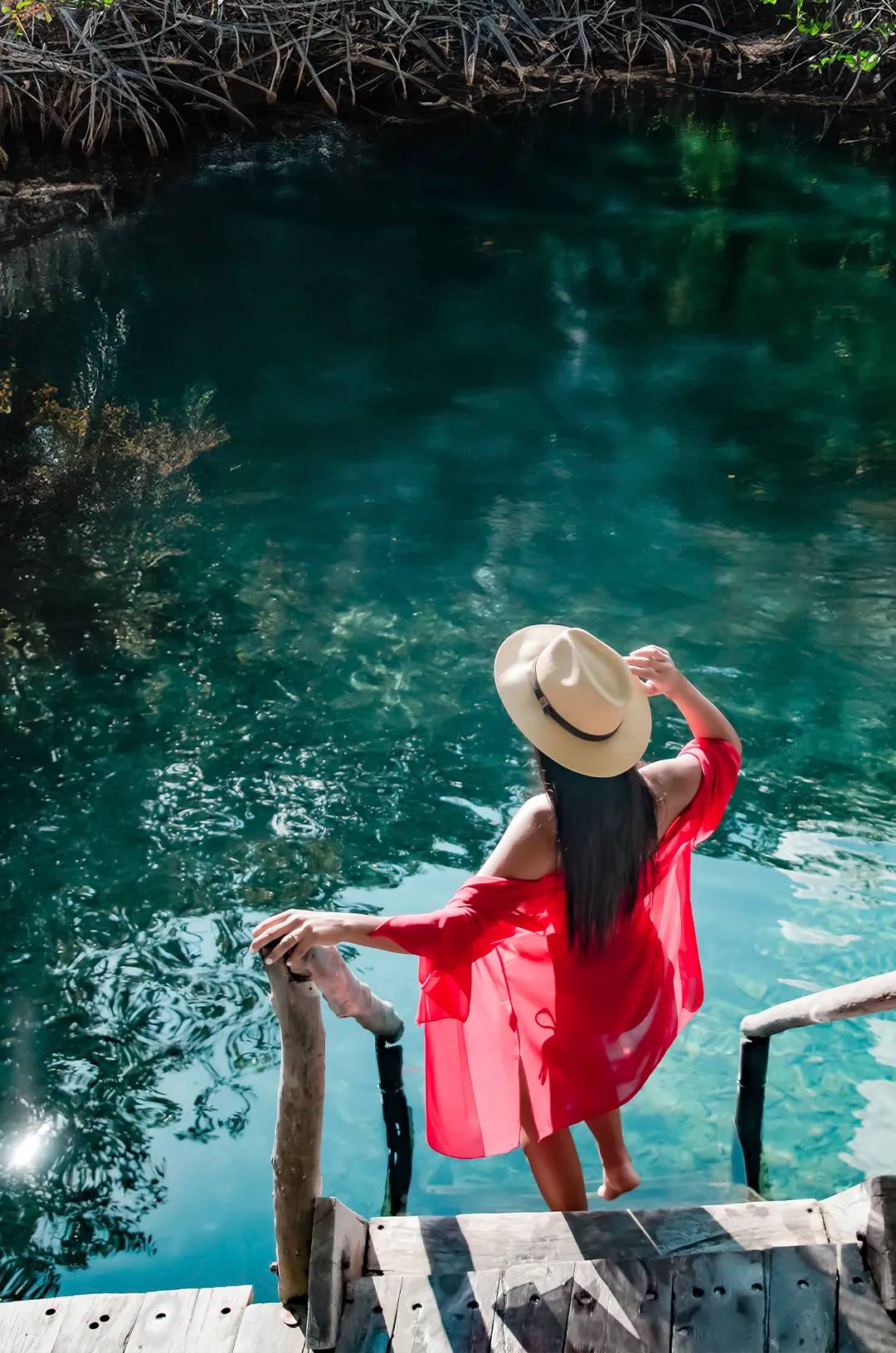 Woman in red dress and hat standing on wooden steps over clear turquoise water