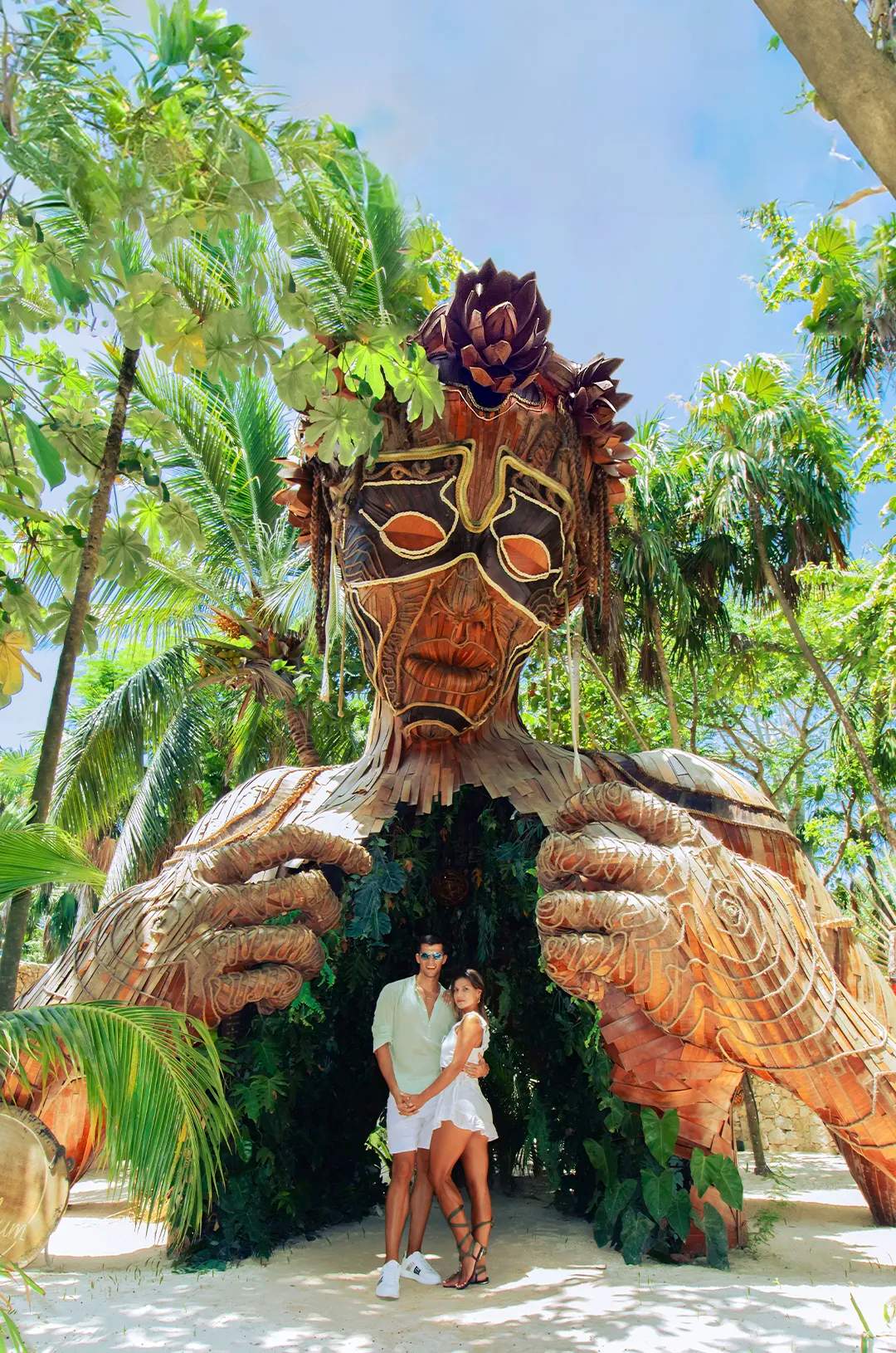 Couple posing in front of the "Ven a la Luz" sculpture in Tulum, surrounded by greenery.
