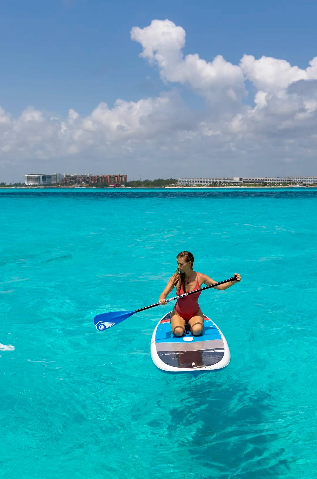 A woman in a red swimsuit kneeling on a paddleboard in turquoise waters, with buildings in the distance.