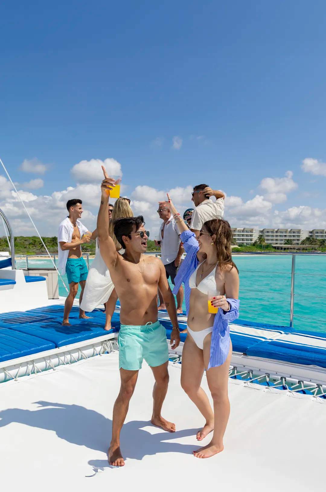 Friends dance and toast with drinks on a catamaran, enjoying the sun, music, and turquoise waters.
