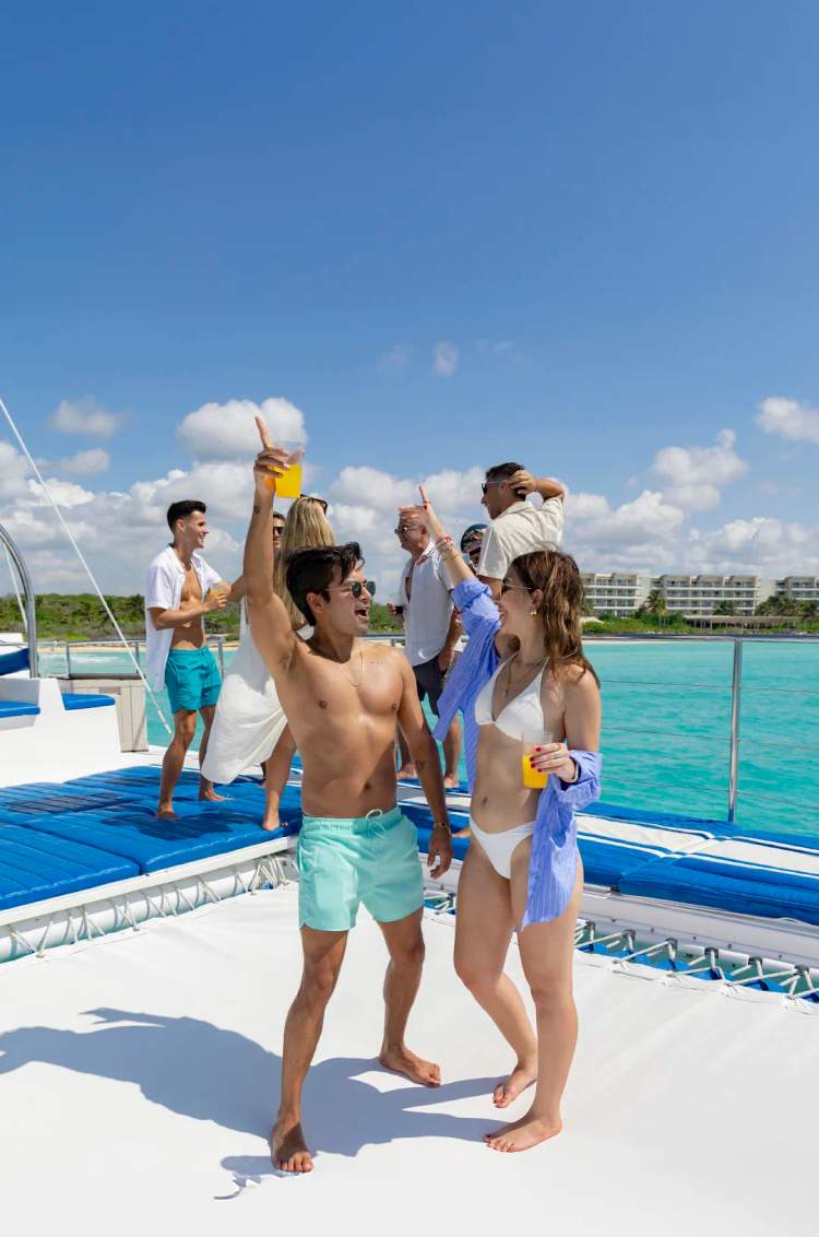 A group of friends enjoys drinks and dances on a catamaran under the sun, celebrating at sea.
