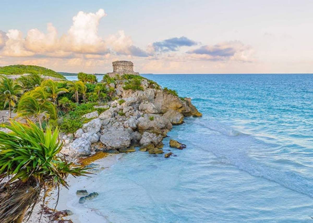 Una vista pintoresca de las antiguas ruinas mayas de Tulum, situadas en un acantilado con vistas a las aguas turquesas del Mar Caribe. Palmeras y terreno rocoso rodean el sitio histórico bajo un cielo parcialmente nublado.
