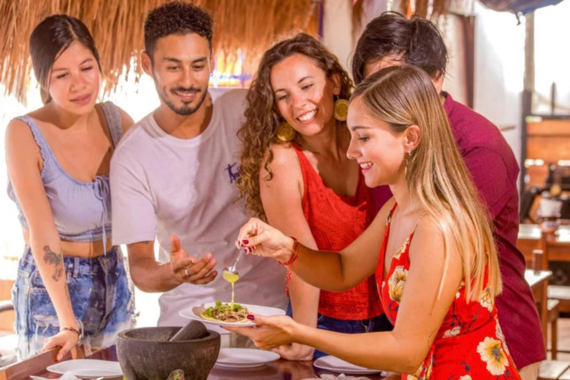 A group of friends enjoying a taco-making experience, smiling and sharing a plate of freshly made tacos with green sauce.