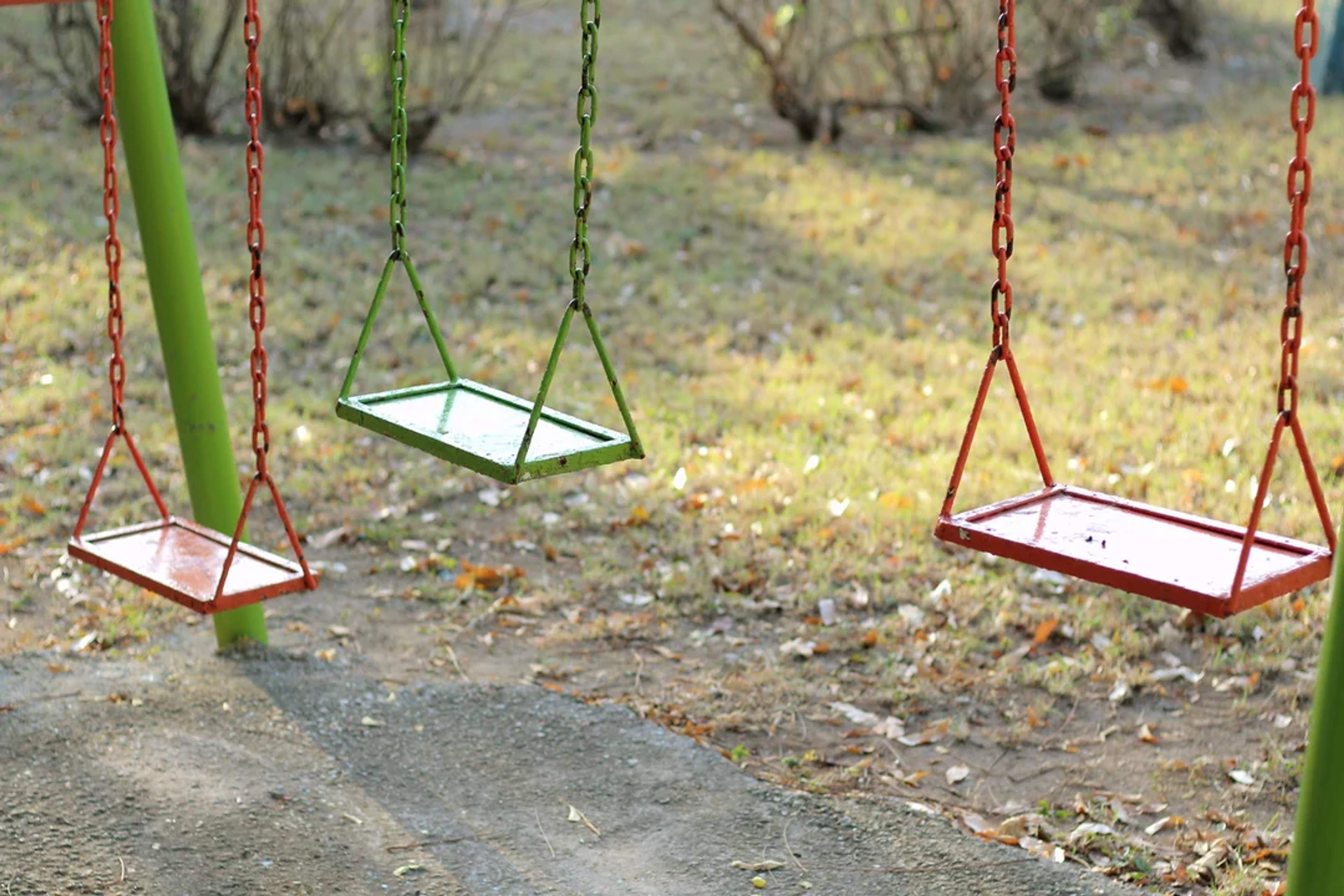 Tres columpios de metal vacíos se balancean suavemente en un parque tranquilo, rodeados de césped y hojas.