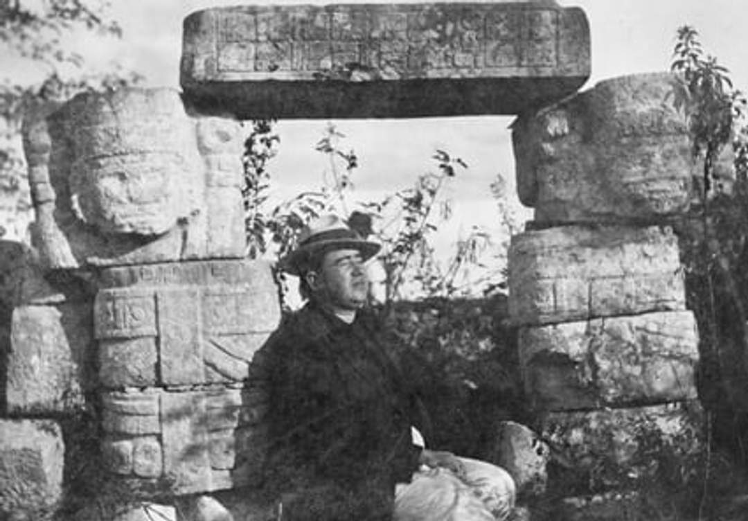 A historical black and white photograph showing a man sitting beneath a stone lintel at Chichen Viejo, surrounded by ancient carved stone structures. The man is wearing a hat and appears to be resting.