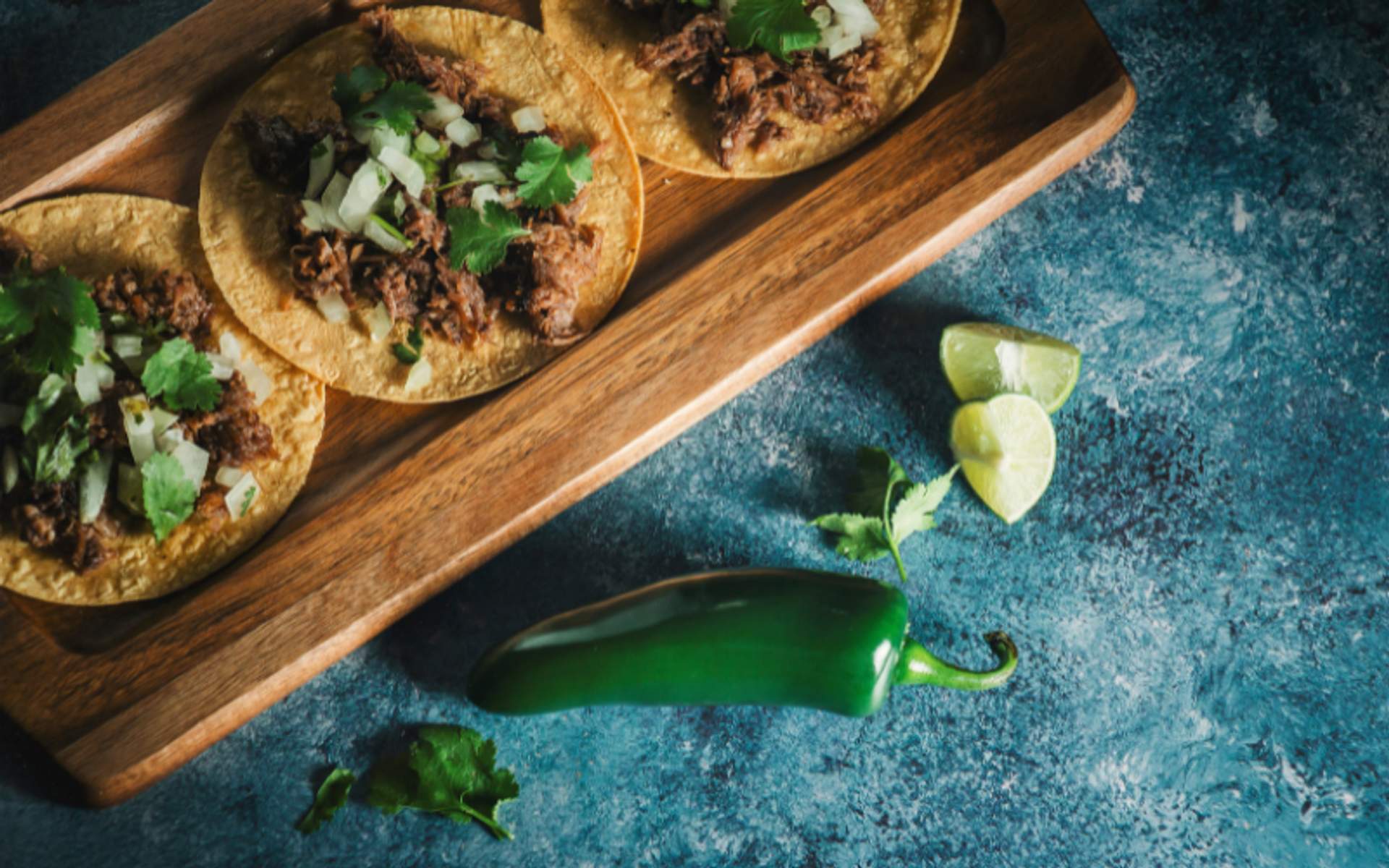 A wooden tray with three tacos topped with cilantro and onions, accompanied by lime wedges and a green chili pepper.