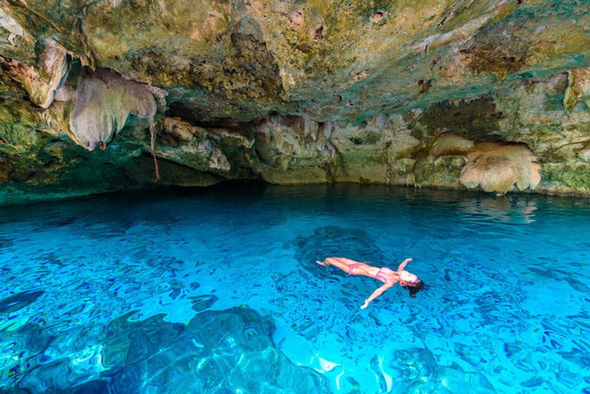 Woman floating in a clear blue cenote, surrounded by rock formations and stalactites in a cave.