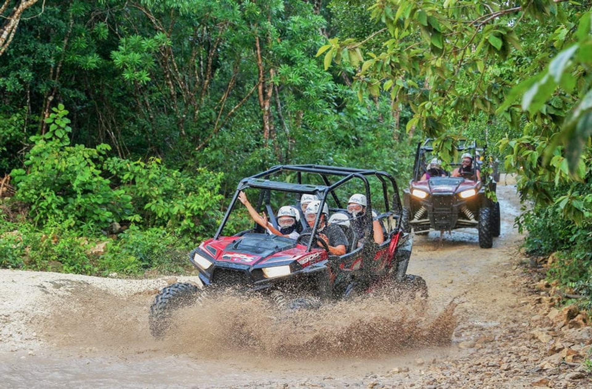 Two all-terrain vehicles splash through a muddy trail in a lush forest, passengers wearing helmets and enjoying the ride.