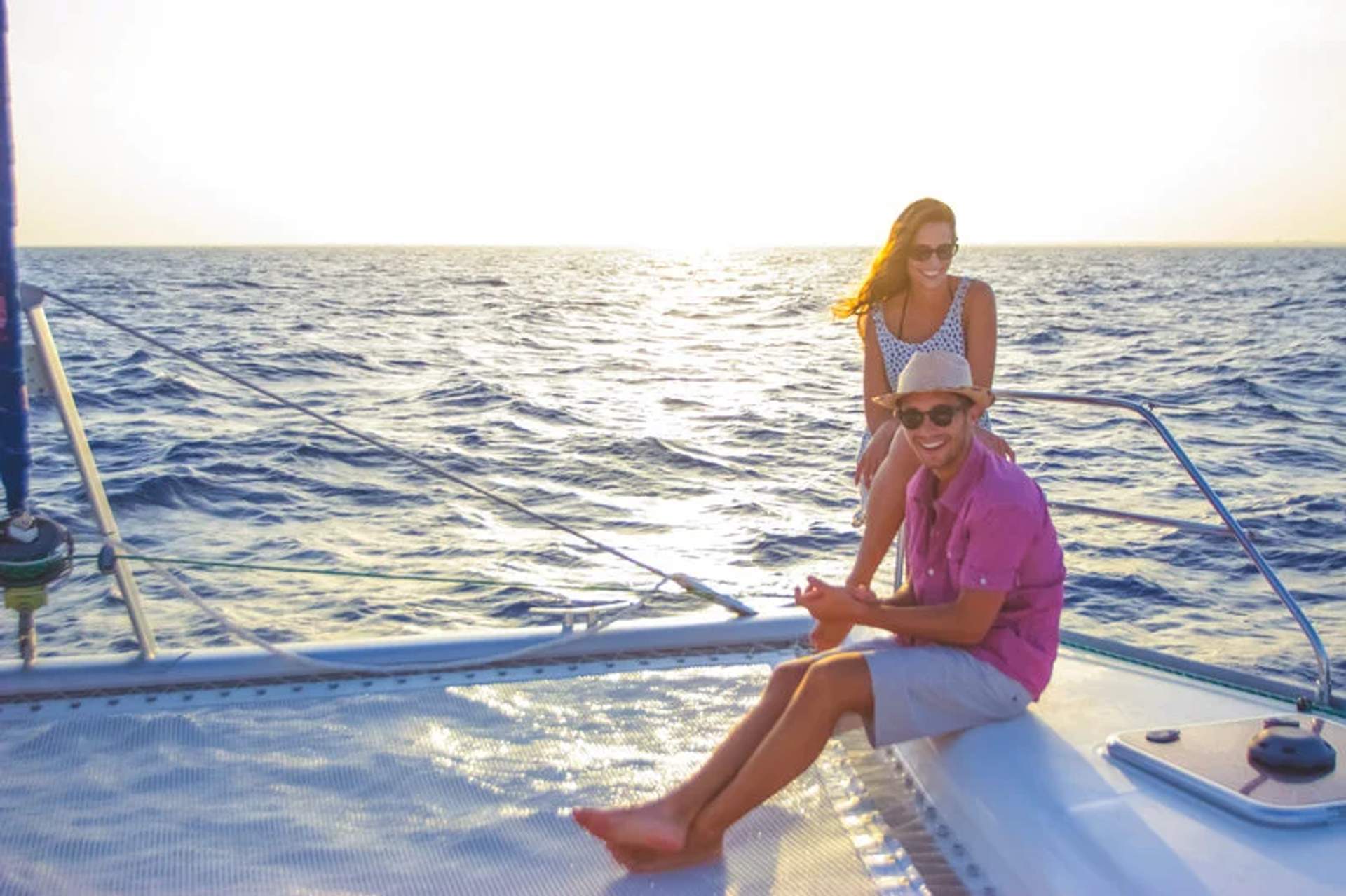 Smiling couple relaxing on a sailboat at sunset, with the ocean and horizon in the background.