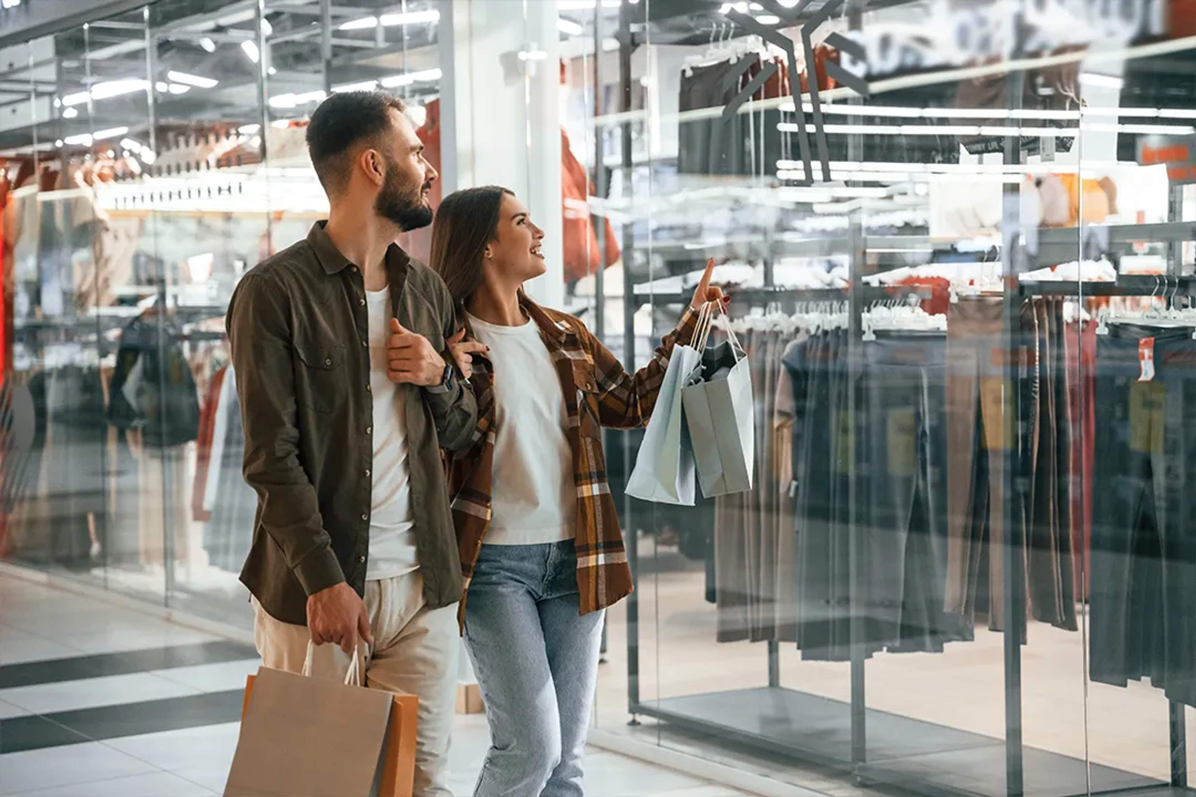 Pareja feliz de compras en un centro comercial, señalando ropa tras un escaparate.