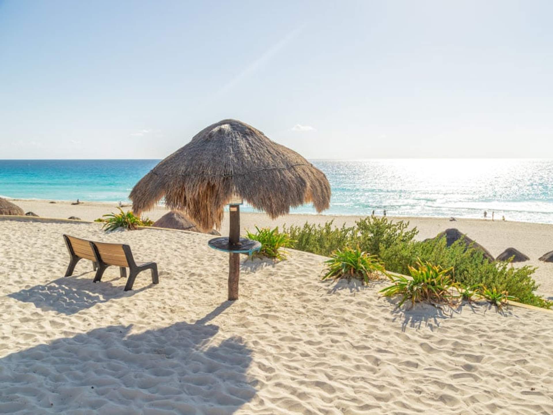 Banco vacío y una sombrilla de paja en una playa de arena con aguas turquesas y un cielo despejado en Cancún.