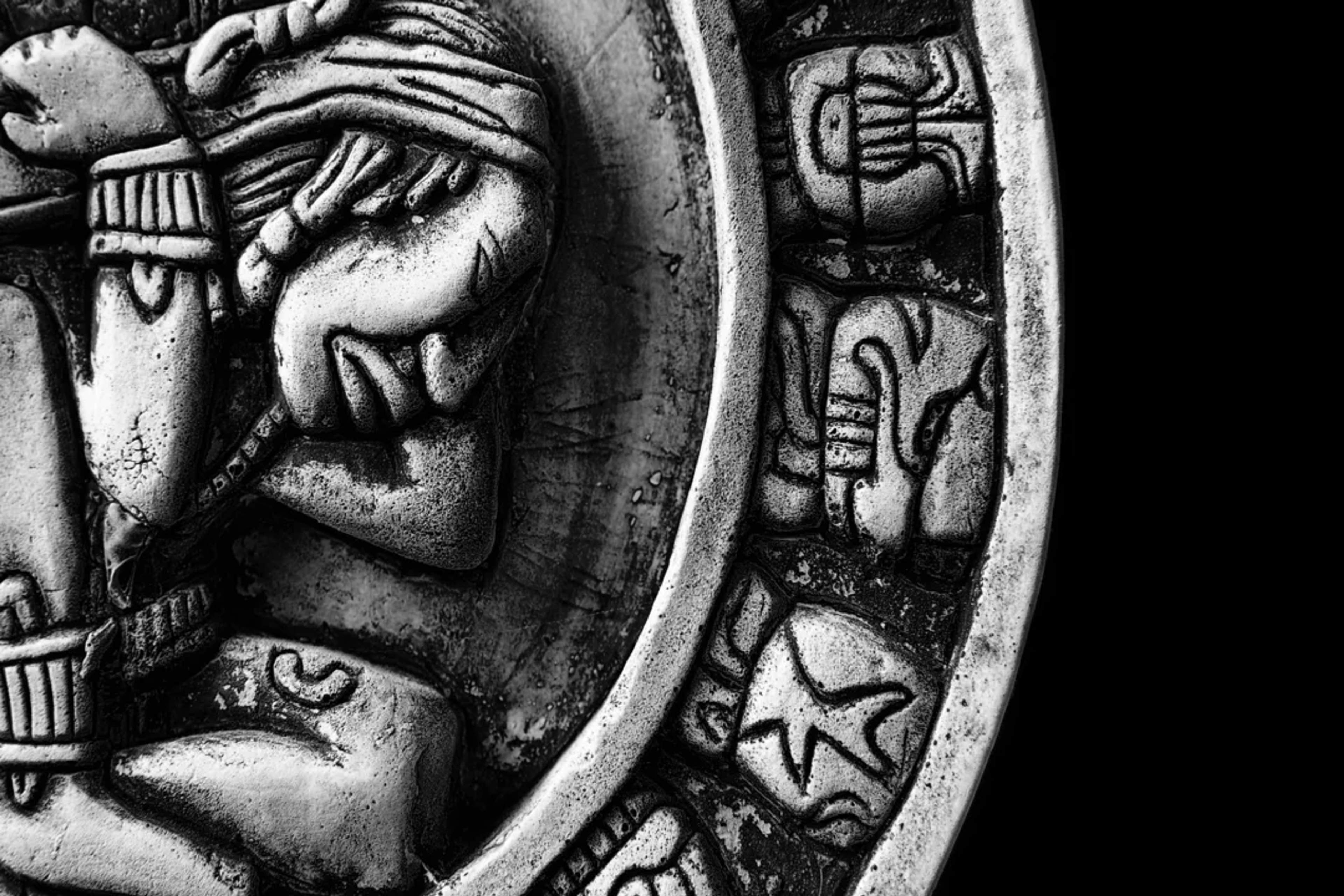 Close-up of a Mayan calendar with intricate glyphs and a sculpted figure in black and white.