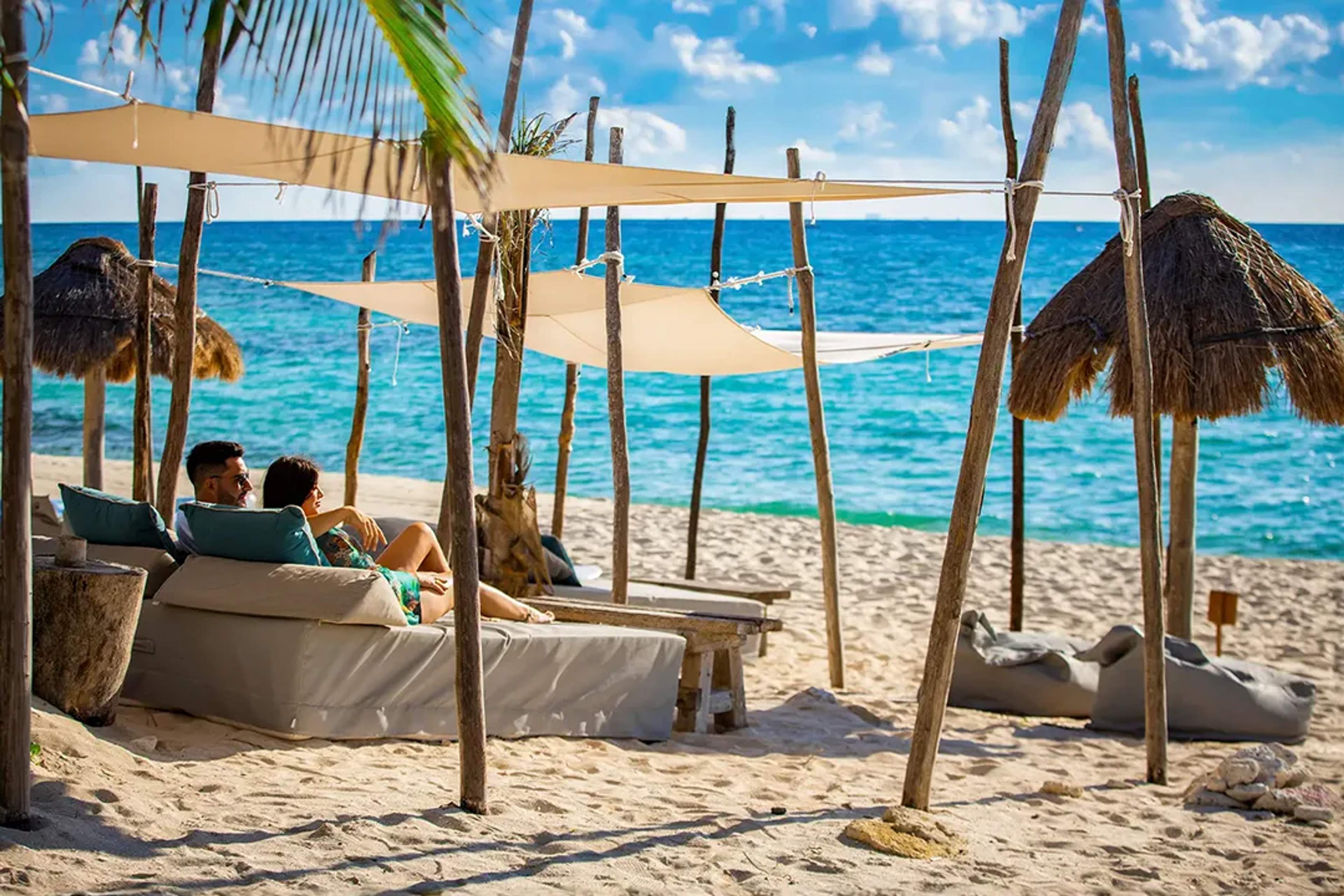Pareja relajándose en una cama de playa con sombra, disfrutando del sol y la brisa marina.