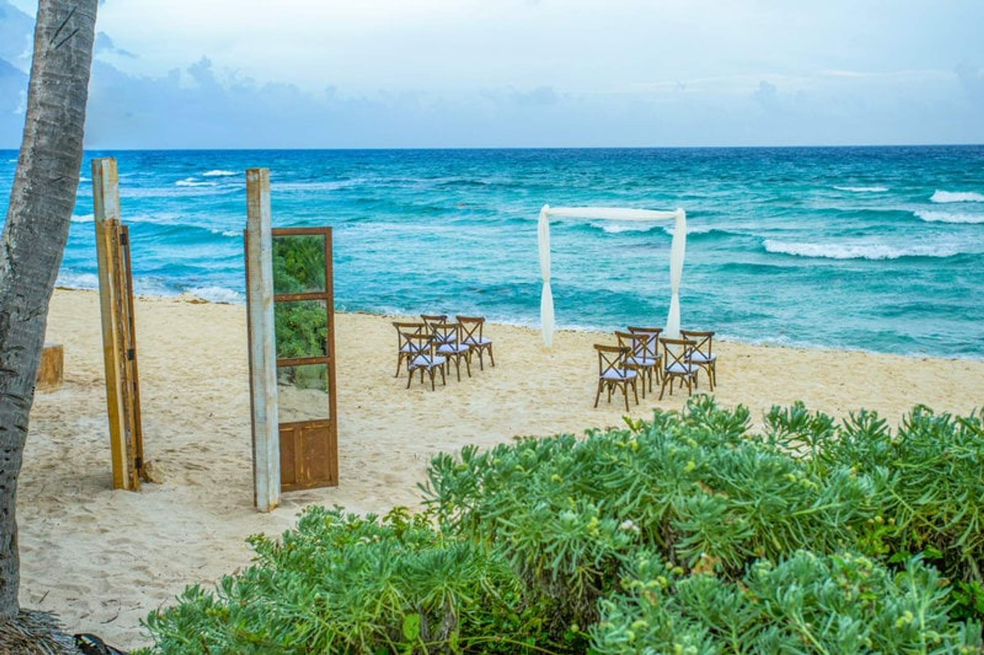 Ceremonia de boda en la playa con sillas de madera dispuestas frente al mar, un arco blanco y puertas decorativas, rodeada de vegetación costera.
