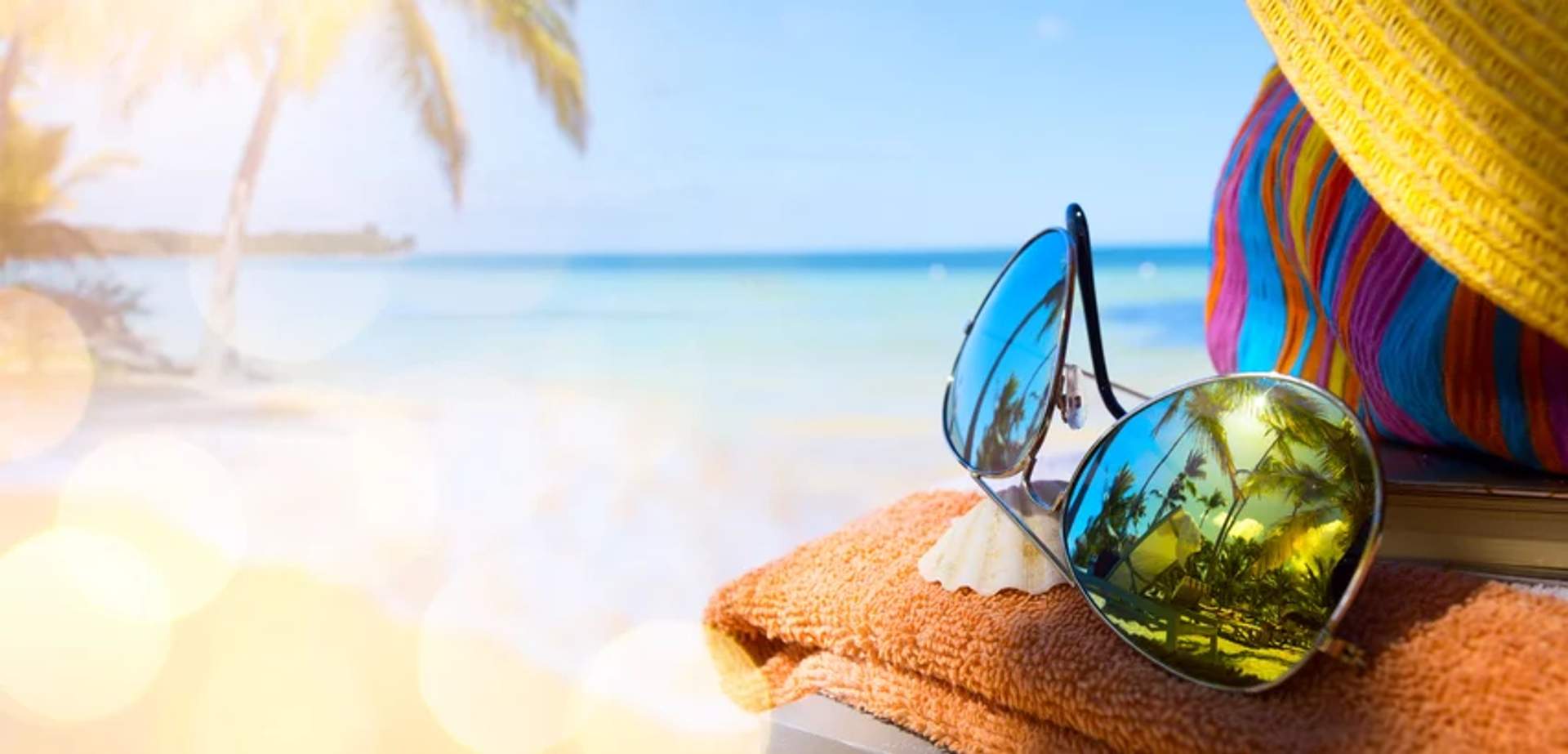 Sunglasses, a shell, and a colorful hat rest on a towel at a sunny beach with palm trees and blue water in the background.