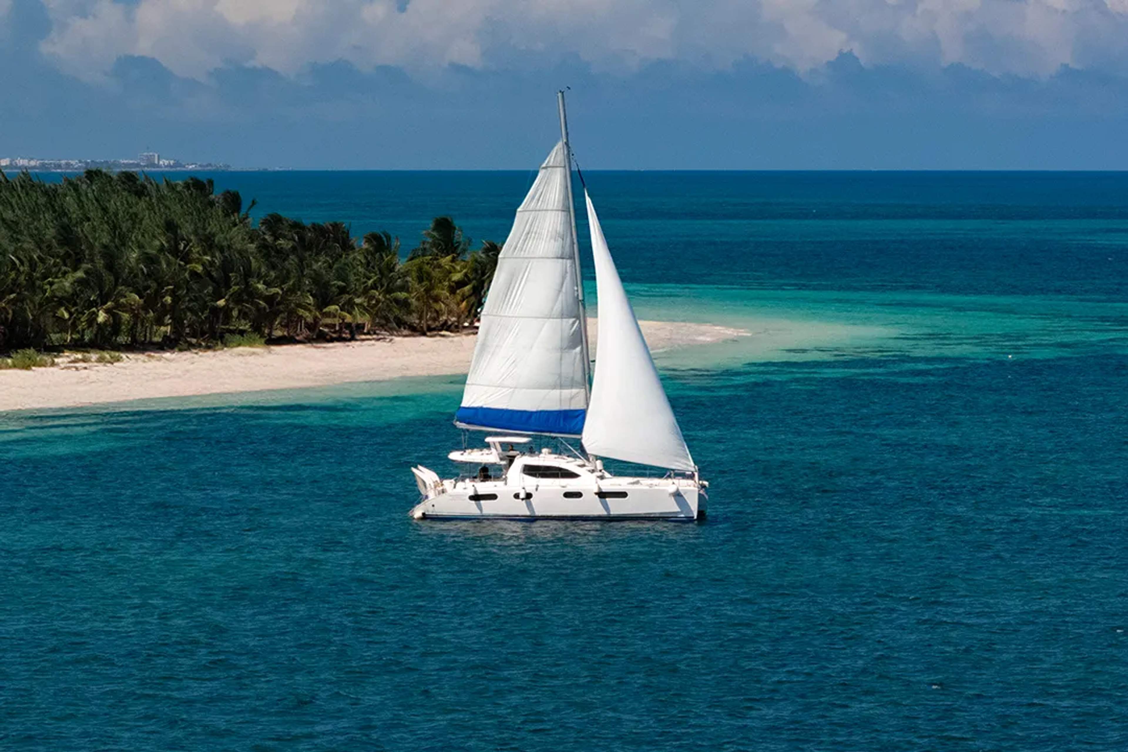 Sailboat gliding over turquoise water near a white-sand island and palm trees in the Mexican Caribbean