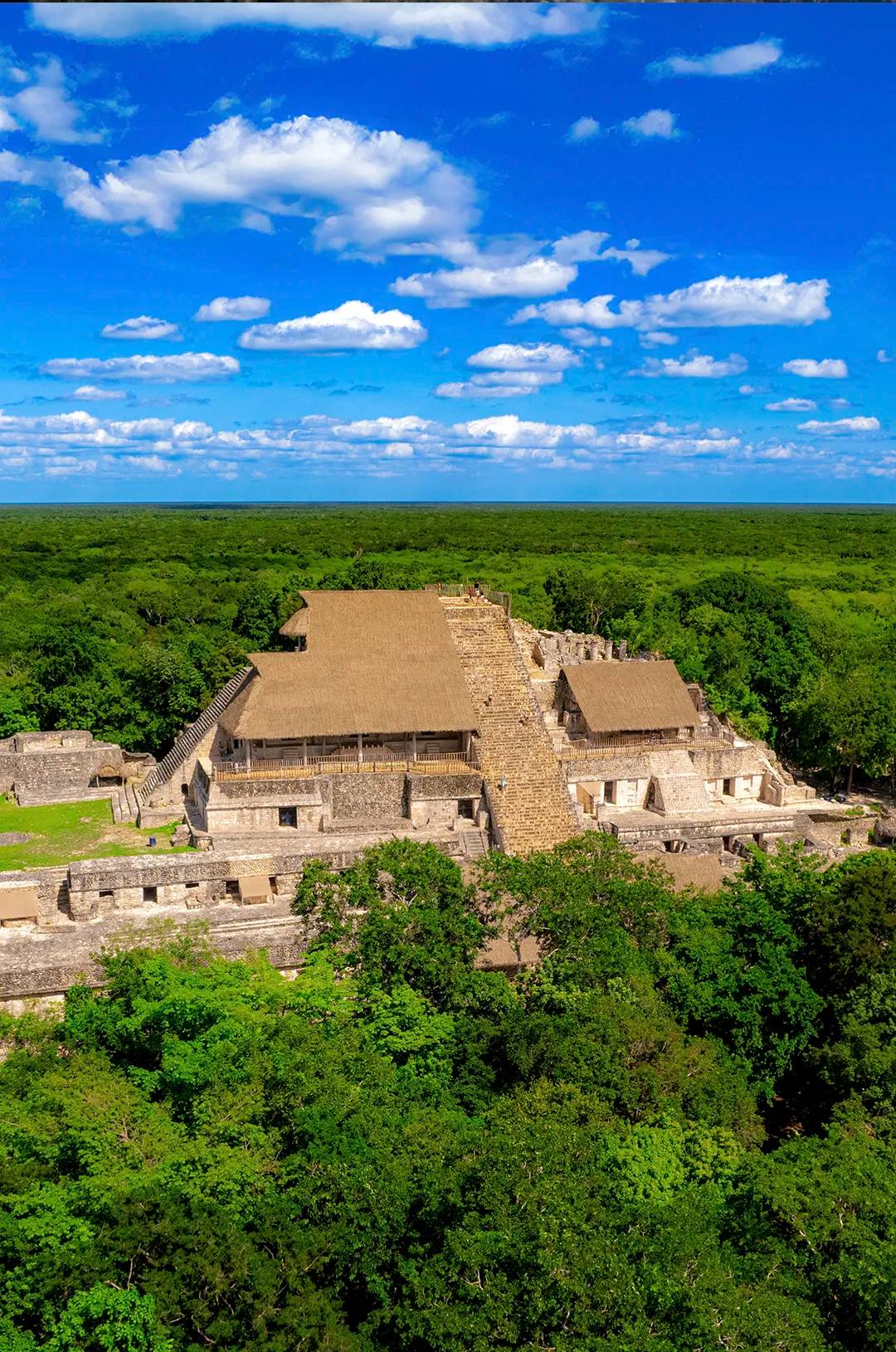 Vista aérea de la Acrópolis de Ek Balam rodeada de exuberante selva y bajo un cielo azul con nubes dispersas