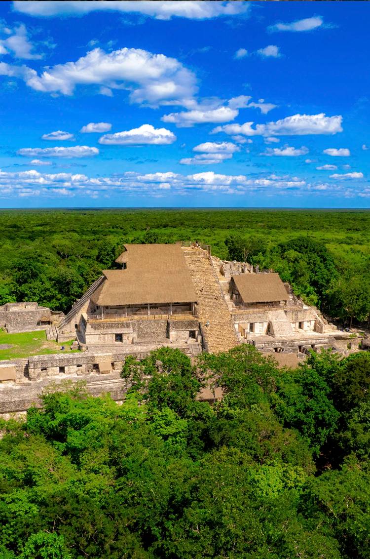 Aerial view of the Acropolis of Ek Balam surrounded by lush jungle and under a blue sky with scattered clouds.