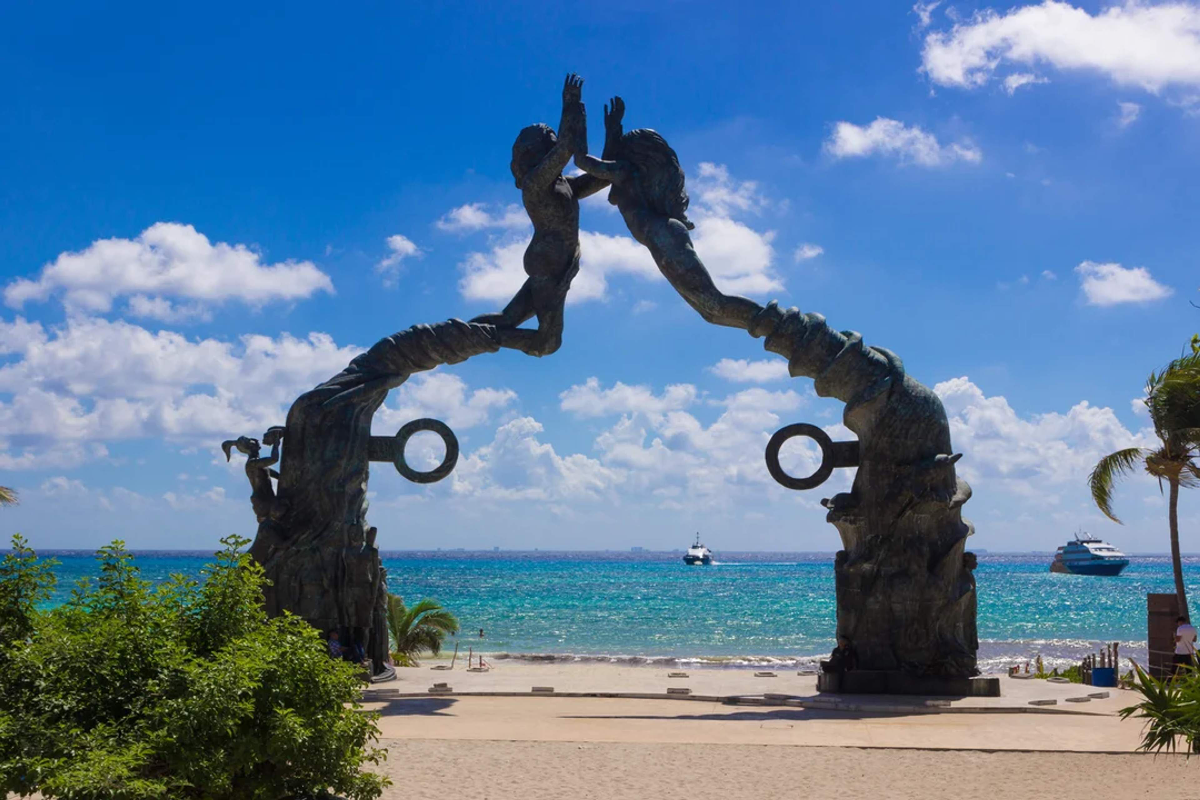 Iconic Playa del Carmen arch sculpture faces the turquoise sea under a bright, sunny sky.