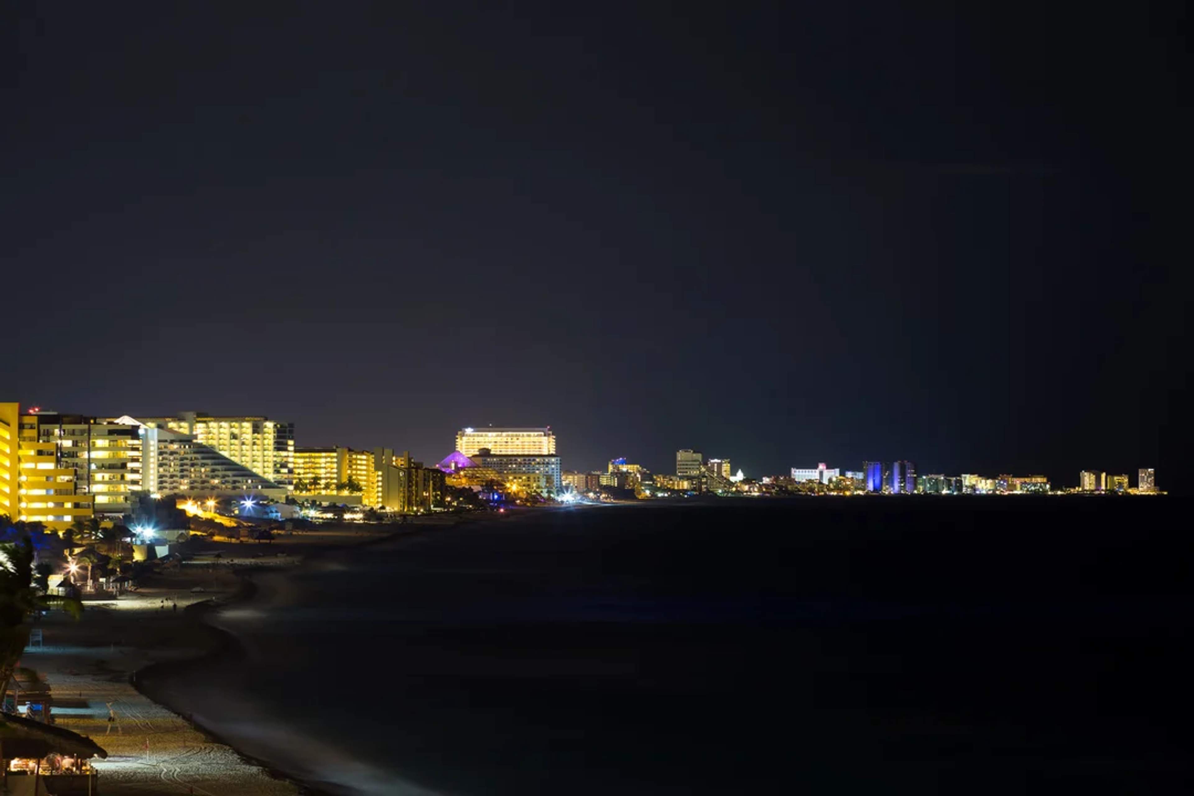 Costa de Cancún de noche, iluminada por luces de hoteles, con el mar y el cielo oscuro al fondo.