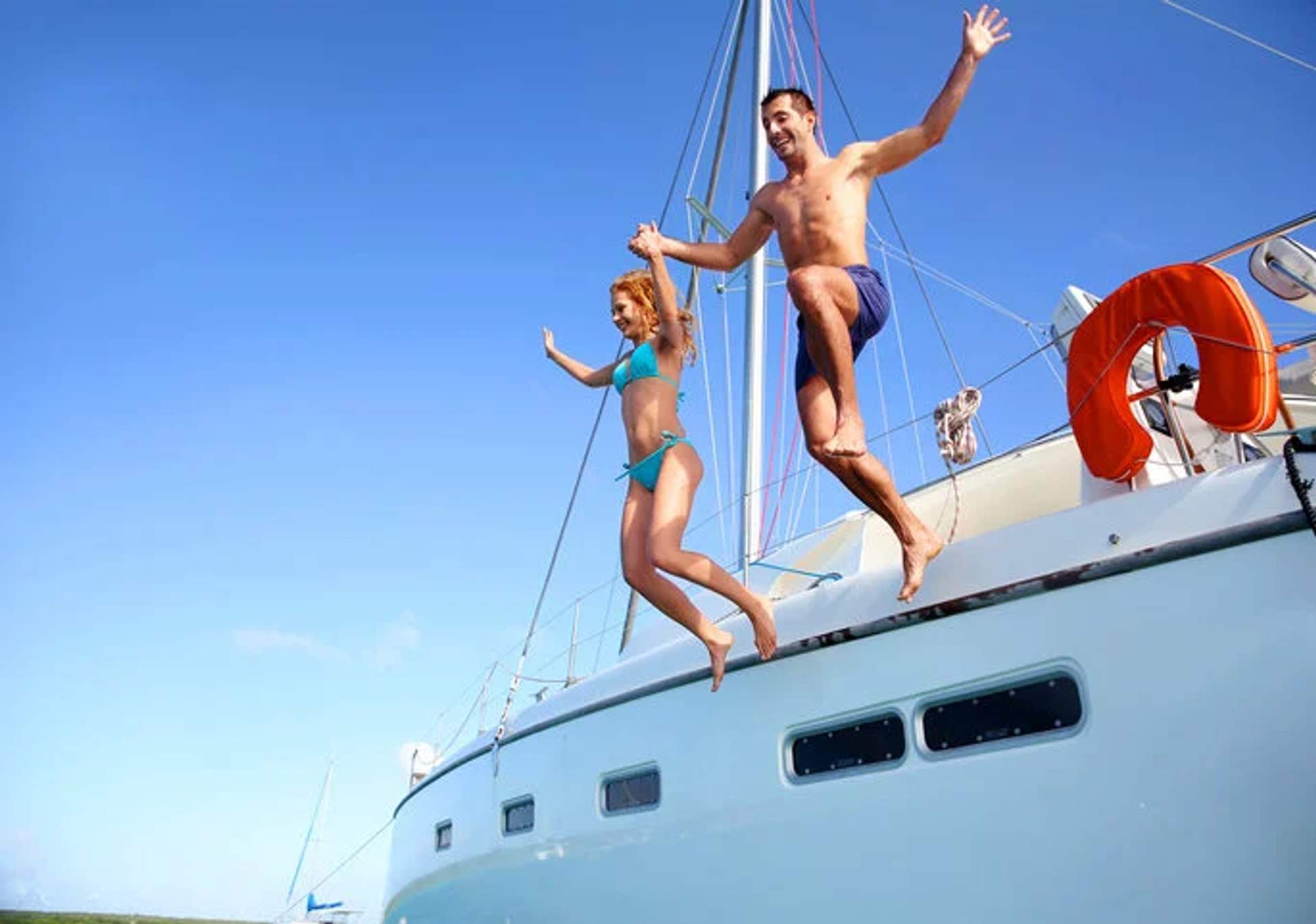 A couple in swimsuits joyfully jumping off a sailboat into the clear blue water on a sunny day.