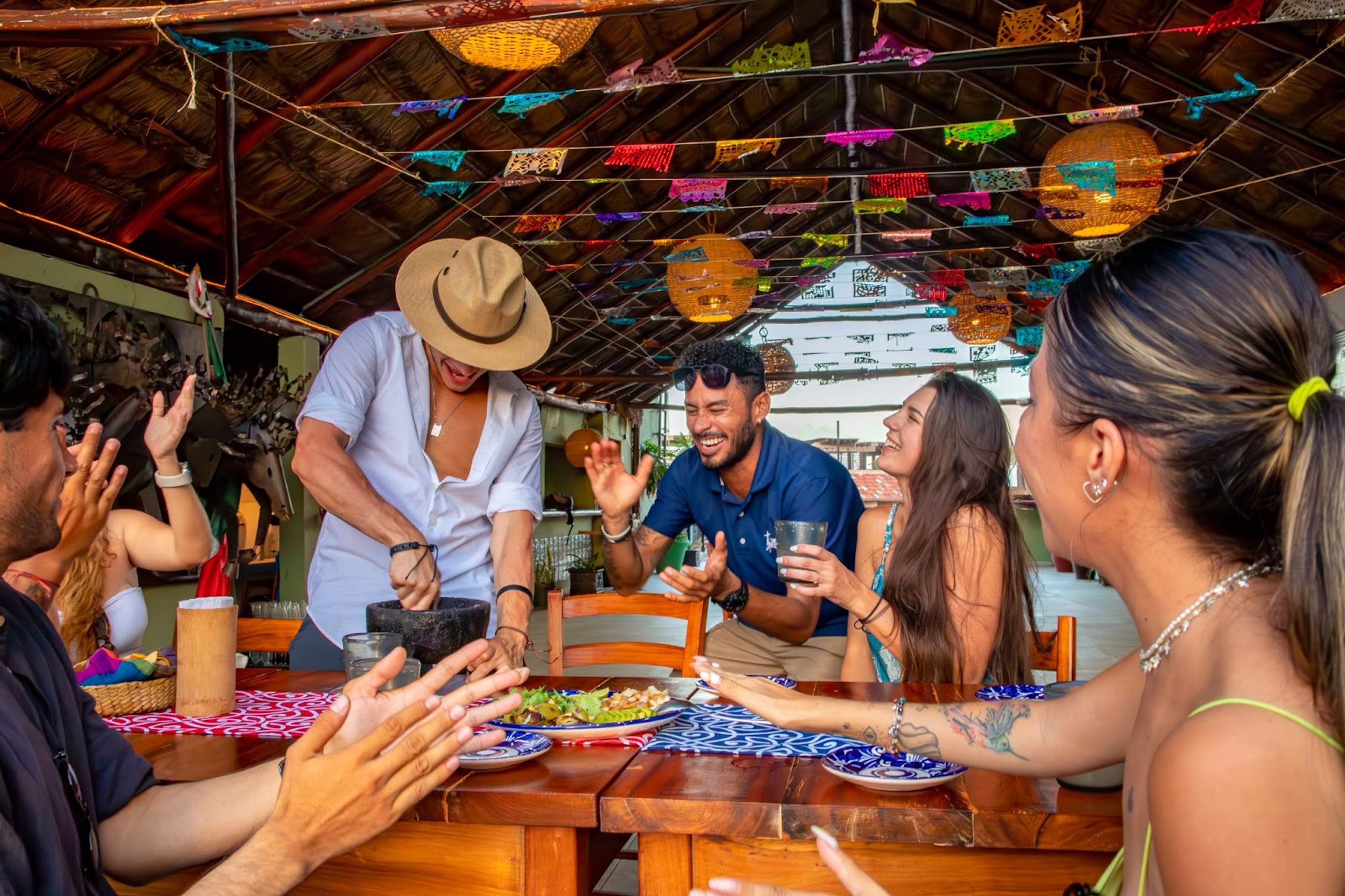 A group of friends laughing and enjoying a meal under a decorated, thatched roof, as one person prepares food with a mortar and pestle.