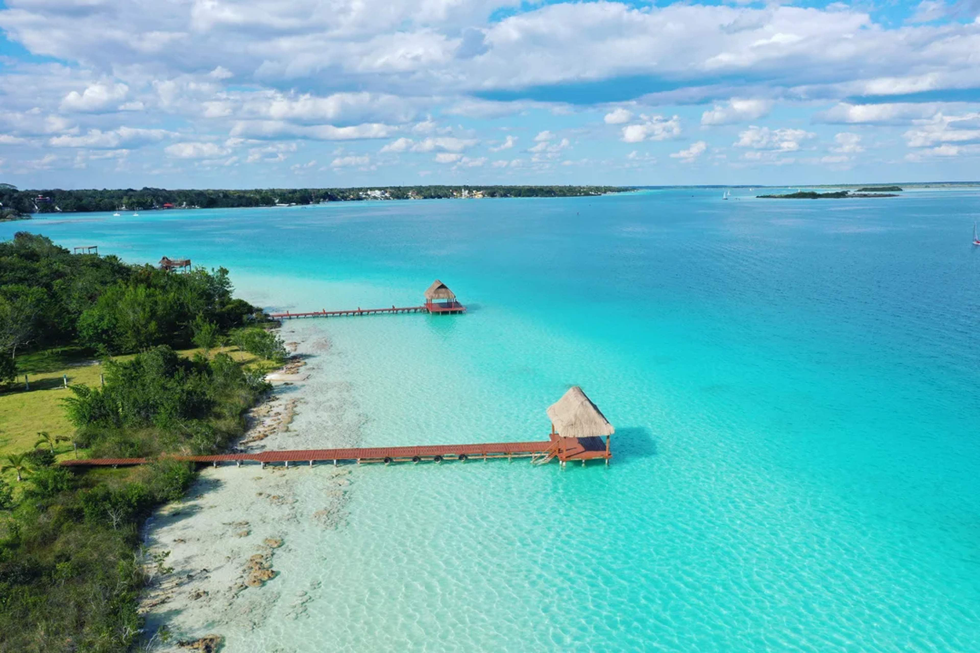 Muelles de madera con palapas se extienden sobre la Laguna de Bacalar, rodeada de aguas turquesas y vegetación exuberante.