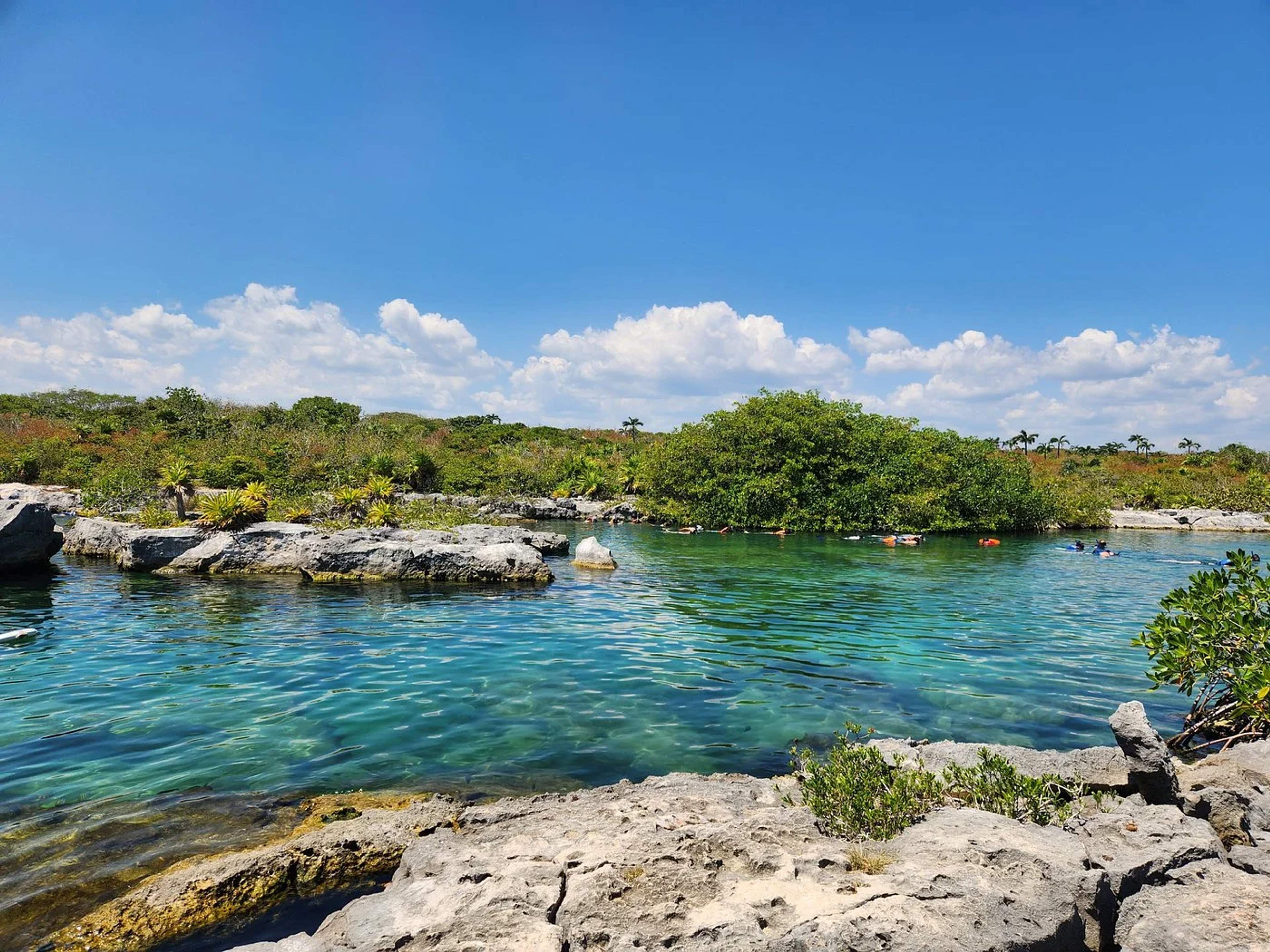 Personas haciendo esnórquel en un cenote de aguas turquesas rodeado de rocas y vegetación exuberante bajo un cielo azul brillante.