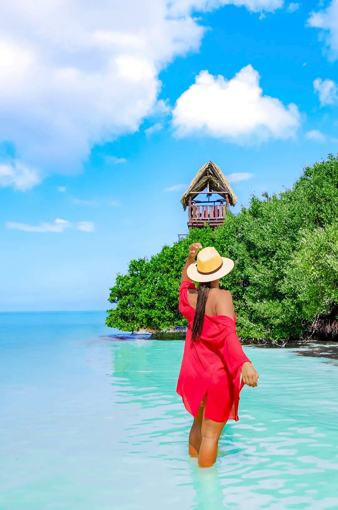 Woman in red dress wading through turquoise water towards a hut