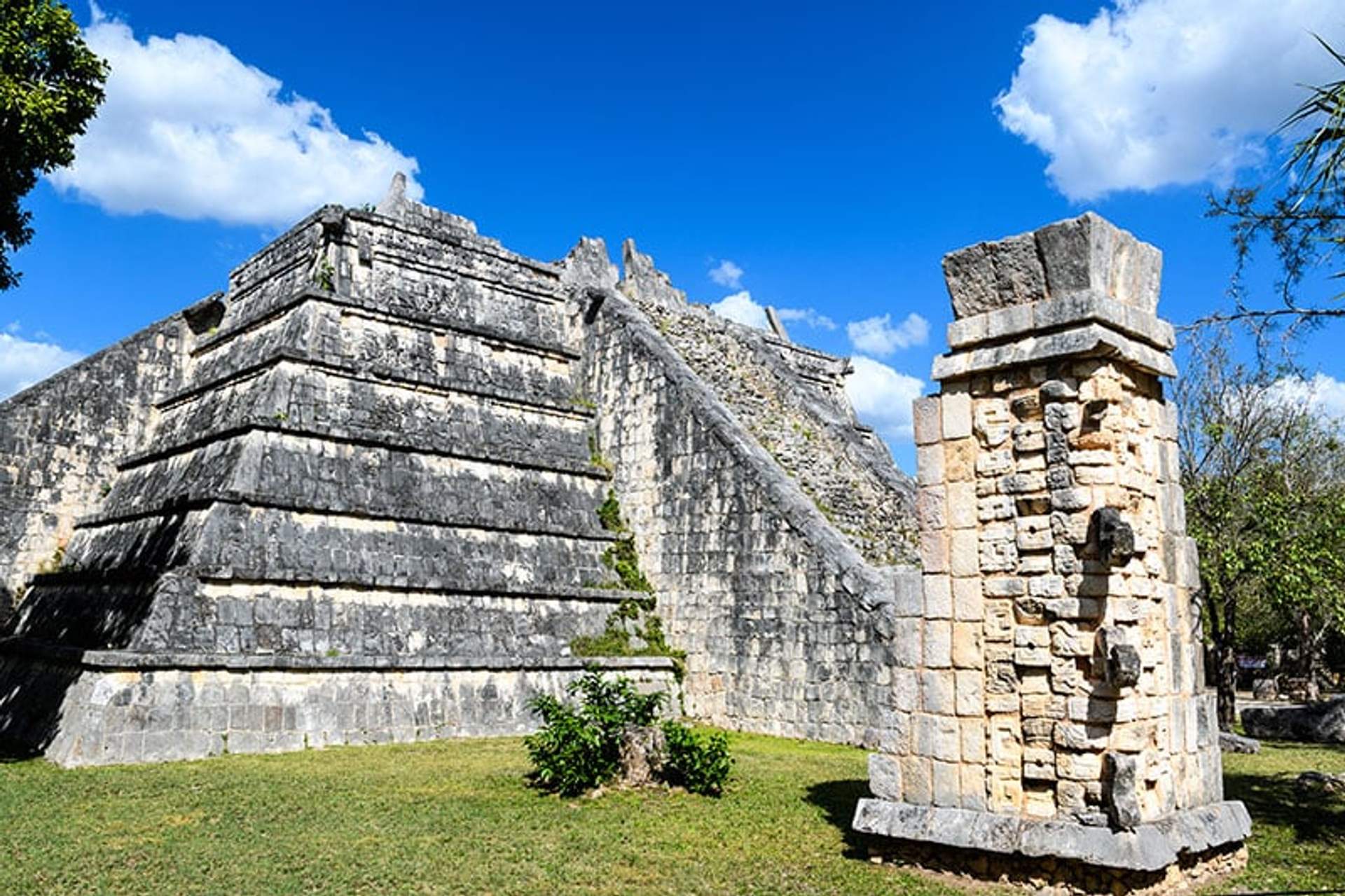 A large stone pyramid structure at an archaeological site under a bright blue sky, with a smaller, weathered stone column in the foreground. The site is surrounded by greenery.