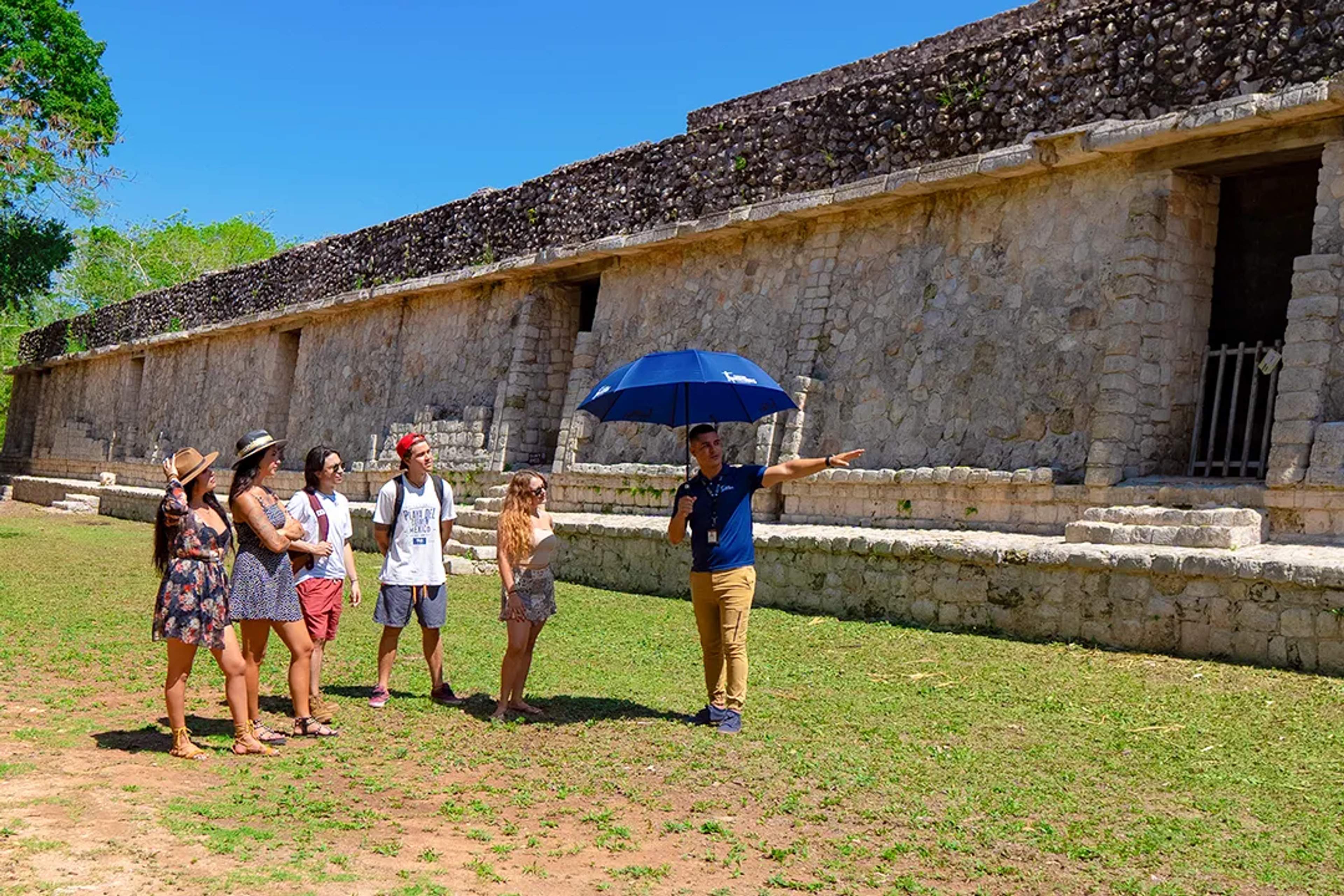 Tour guide explains Mayan ruins to a group of tourists under the sun with an umbrella for shade.