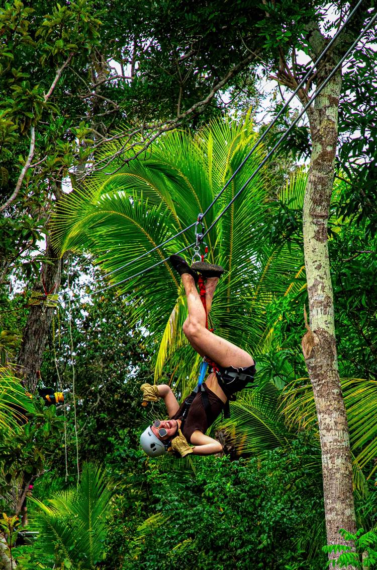 A woman hanging upside down on a zipline, smiling, and surrounded by lush green foliage.