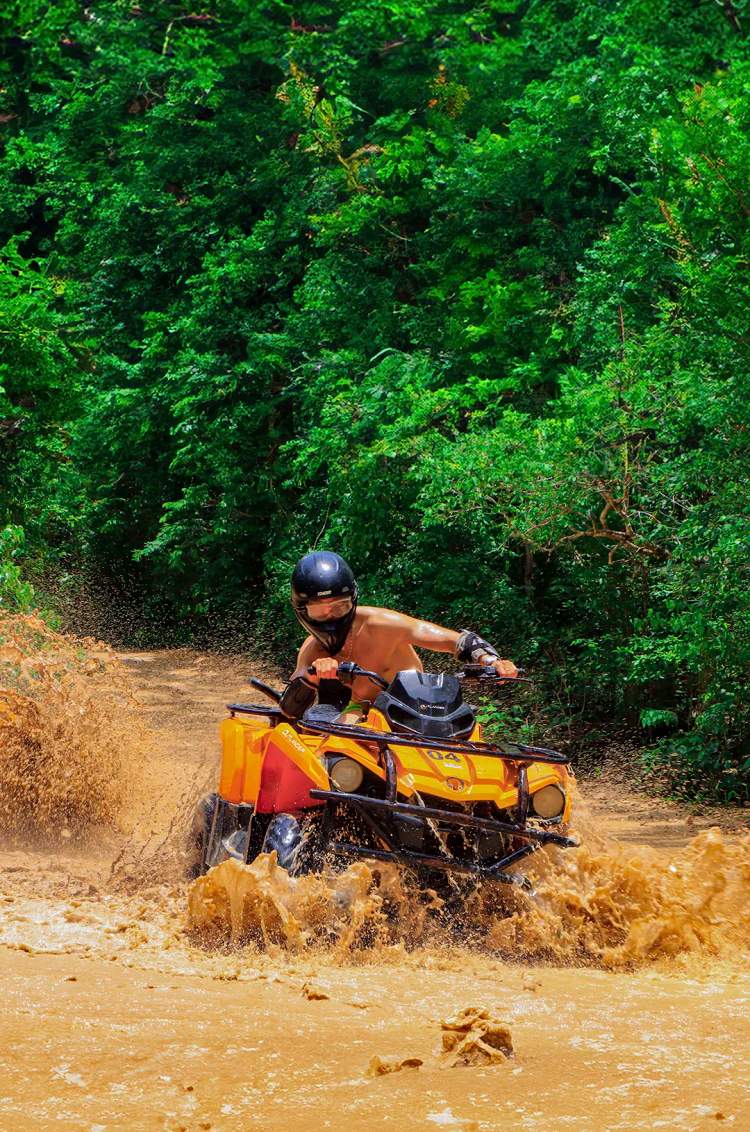 A person riding an ATV through a muddy puddle on a forest trail, splashing water everywhere, wearing a helmet.