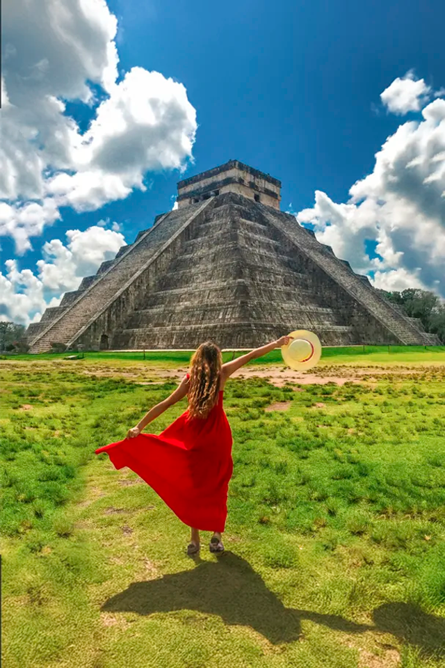 Mujer con vestido rojo y sombrero caminando hacia la pirámide de Chichén Itzá bajo un cielo azul vibrante con nubes.