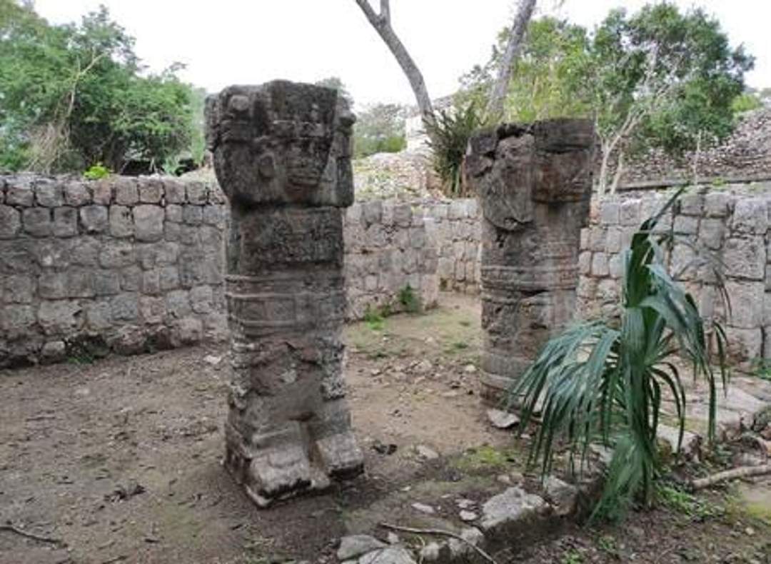 A photograph of two carved stone columns at the House of the Phallus in Chichen Itza, surrounded by partially preserved stone walls. The columns feature intricate carvings, and the site is enveloped by greenery. Photo credit: Giovanni Agostino Frassetto.