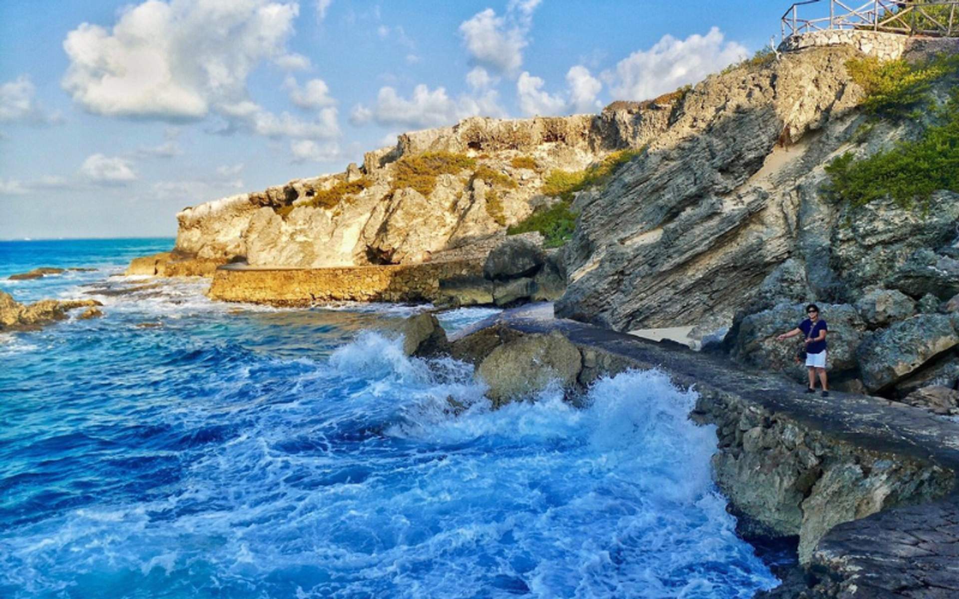 A person standing on a rocky path by the crashing waves at Isla Mujeres, with cliffs in the background.