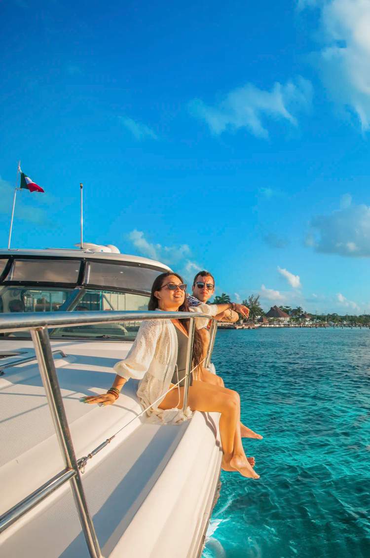 Couple enjoying the sun on the bow of a yacht, overlooking the turquoise sea and Cozumel's coastline.