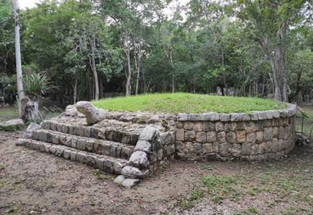 A photograph of the Platform of the Turtle at Chichen Itza, featuring a circular stone platform with a small staircase leading up to it. The platform is surrounded by trees and greenery.
