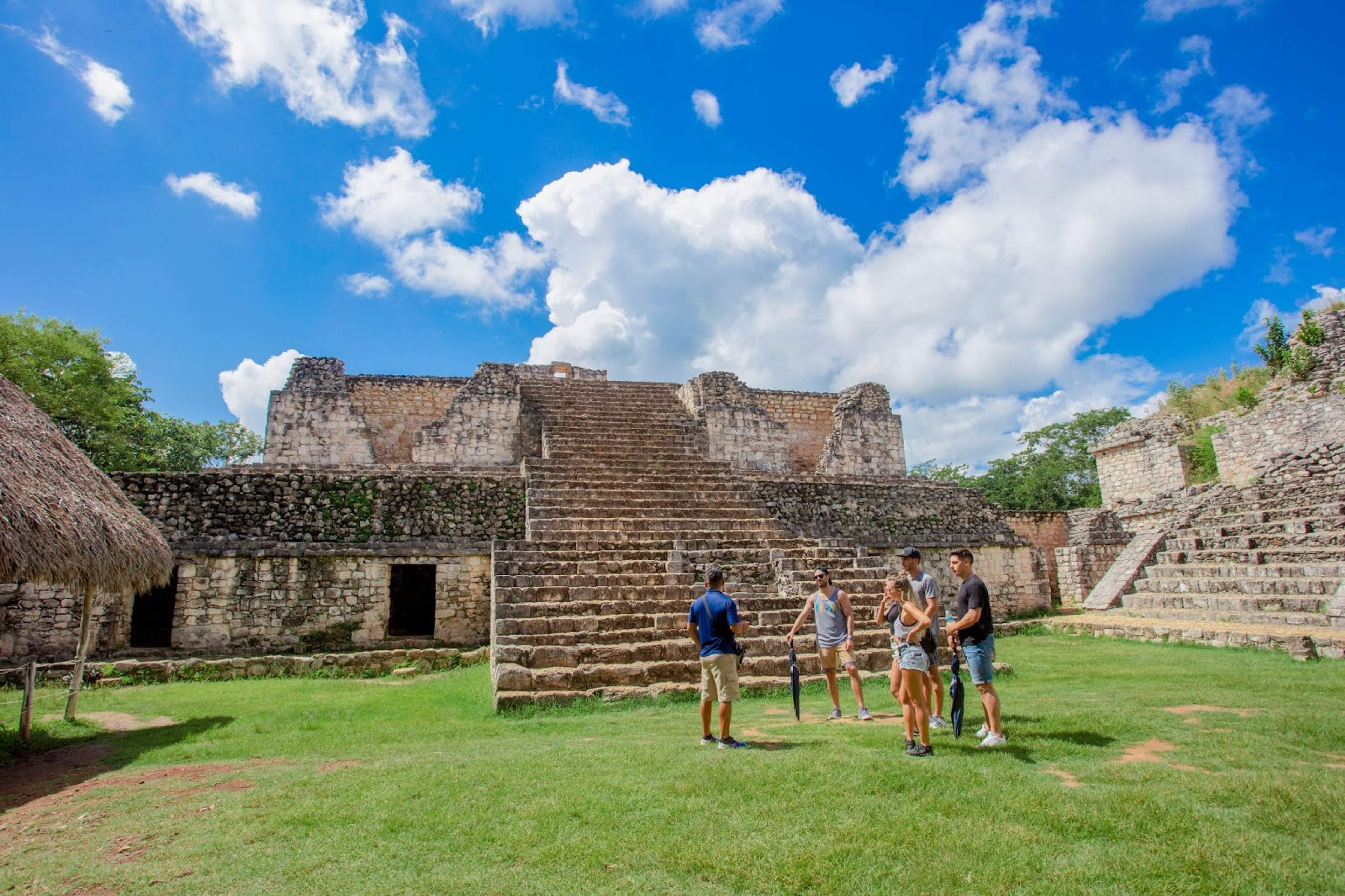 Tourists exploring ancient Mayan ruins with a stone staircase under a bright blue sky with fluffy clouds.