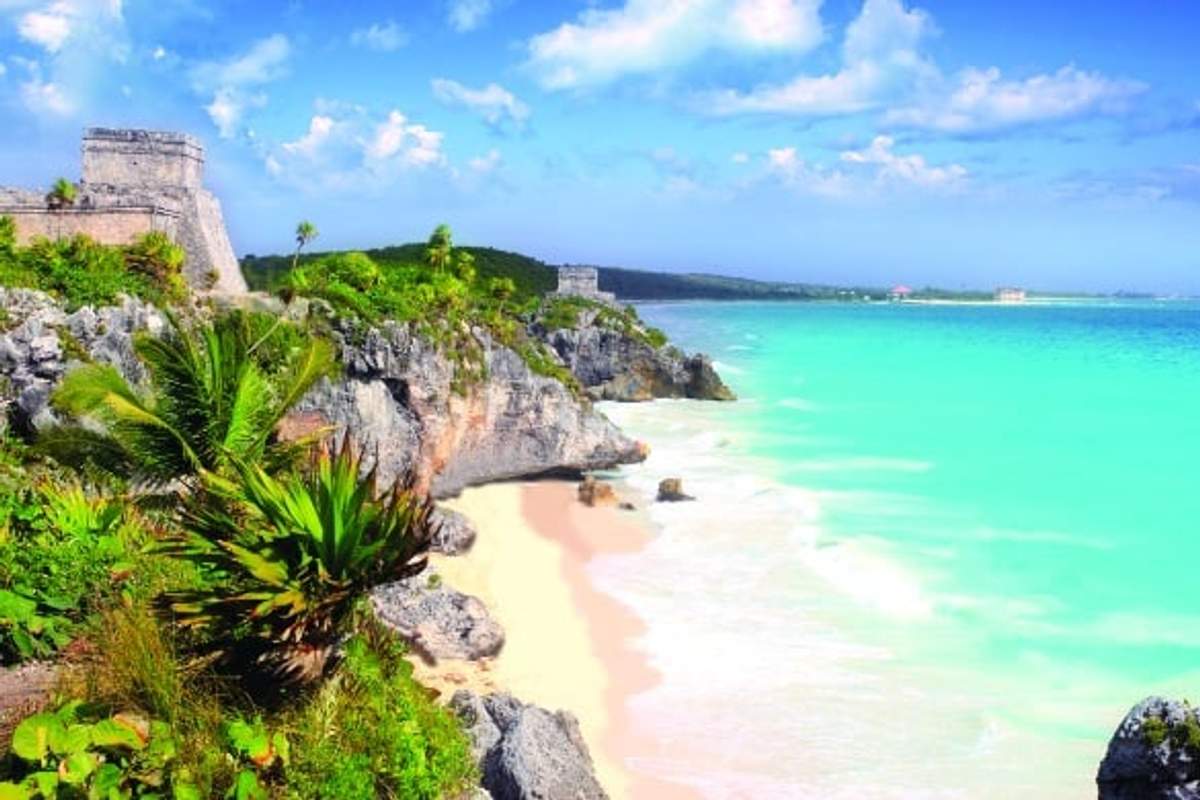 Stunning view of Tulum ruins perched on cliffs overlooking a turquoise sea with lush greenery and a sandy beach below.