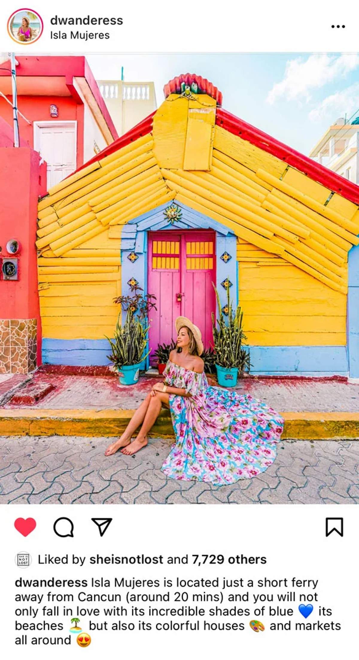 Una mujer con un vestido floral y sombrero de paja sentada frente a una casa colorida amarilla y roja con una puerta rosa en Isla Mujeres.
