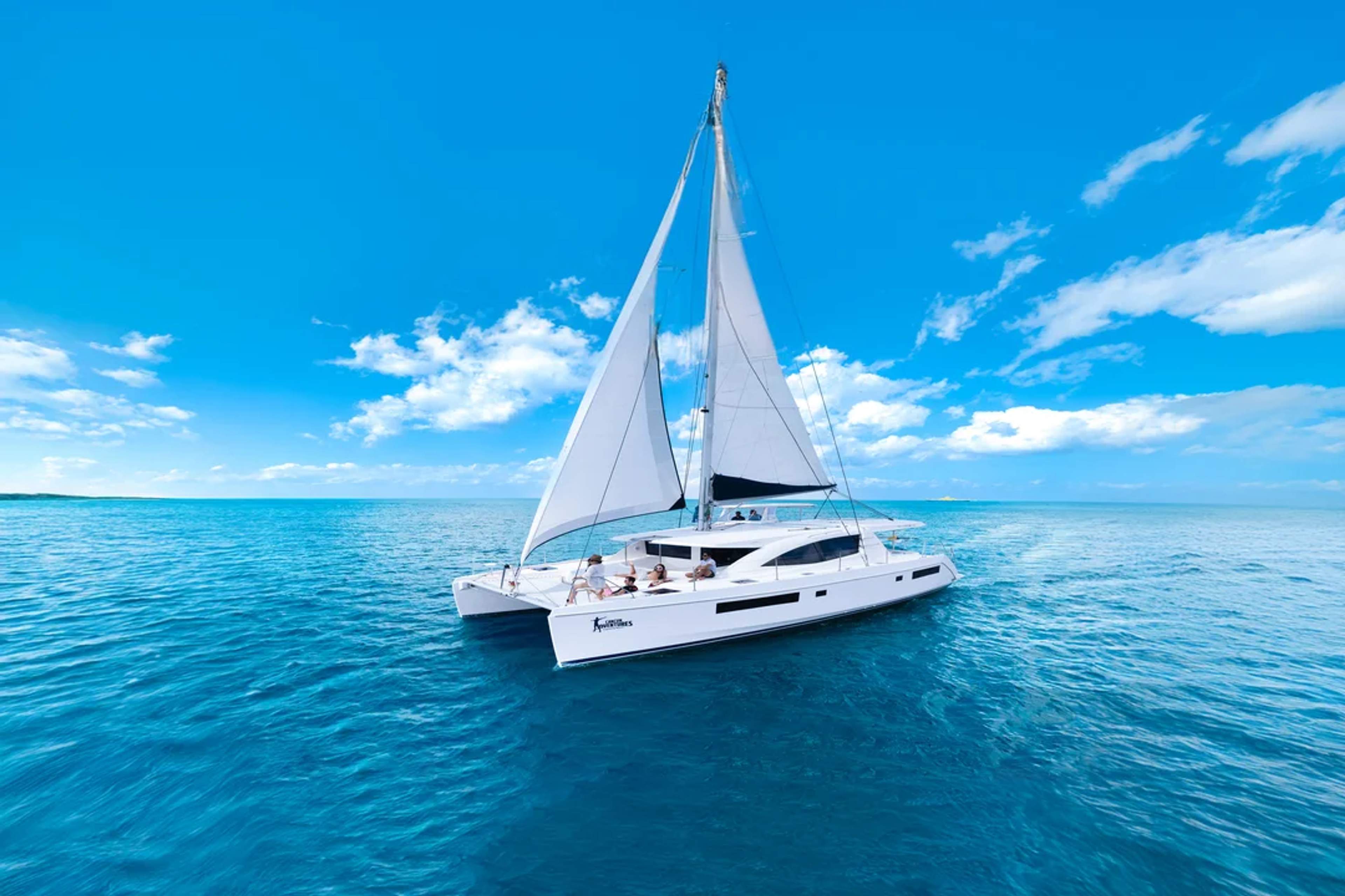 Un catamarán navega por aguas cristalinas bajo un cielo soleado, cerca de una costa rocosa con vegetación exuberante.