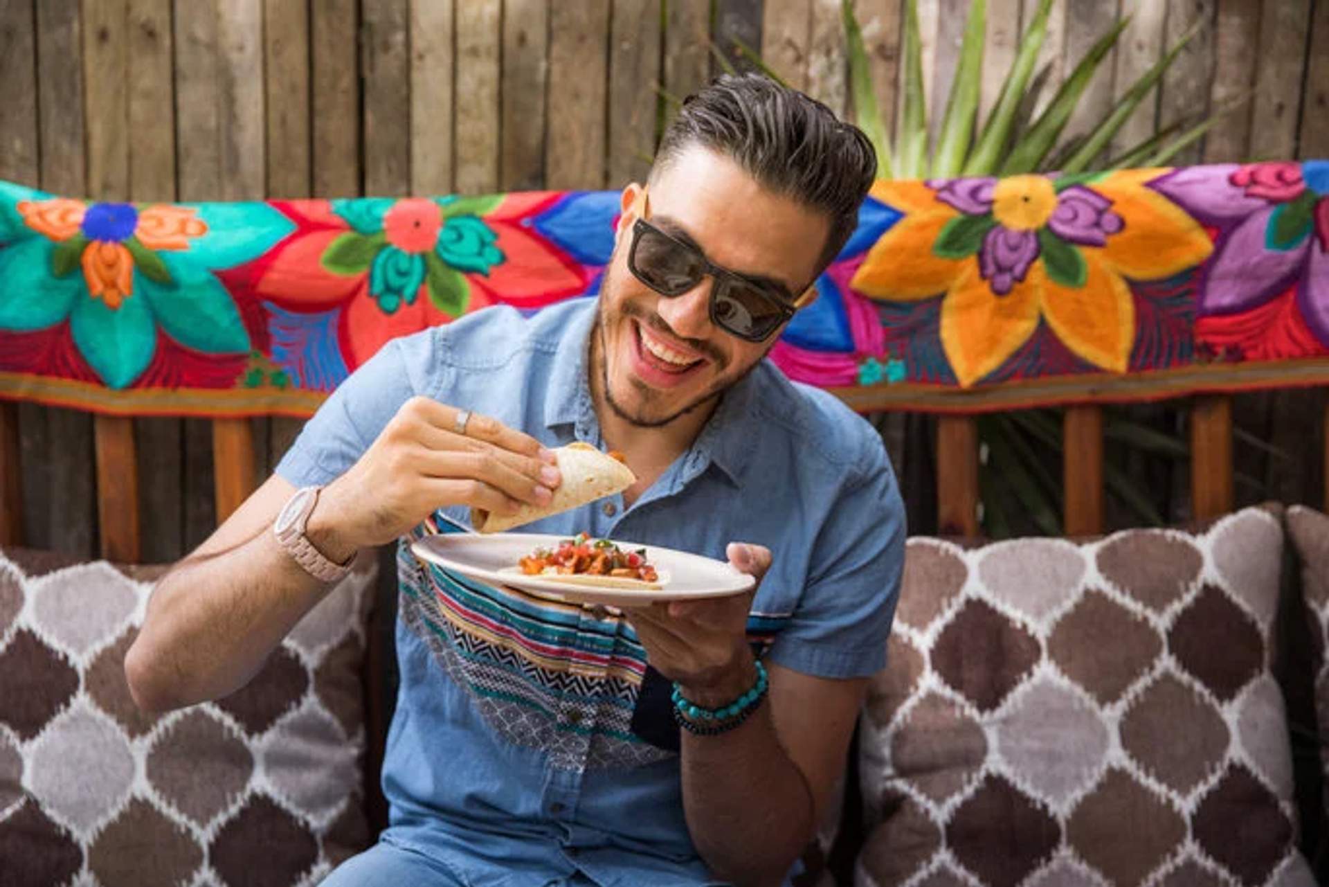 A man wearing sunglasses and a blue shirt smiles while enjoying a taco, sitting on a colorful bench with Mexican decorations.