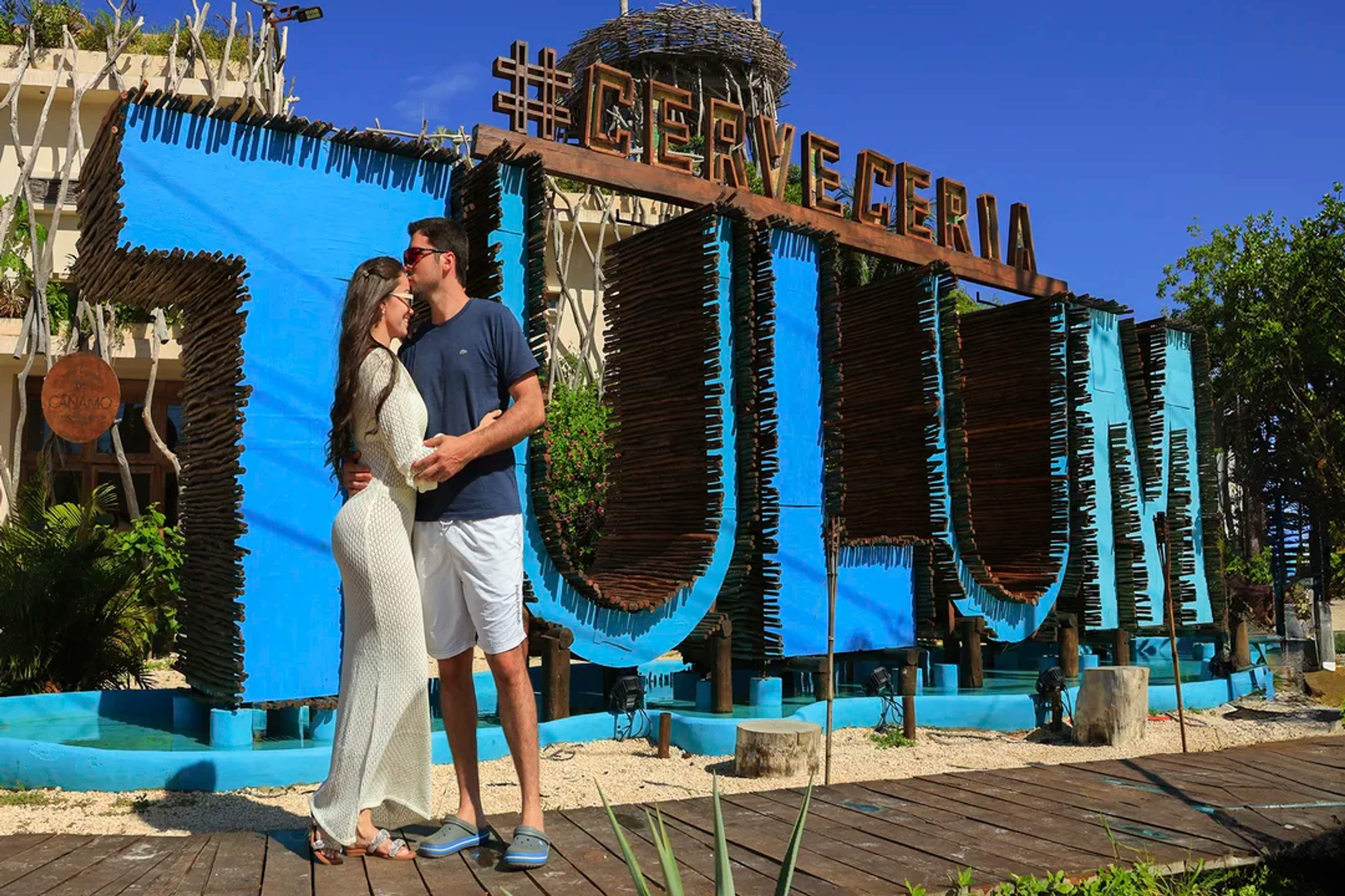 Pareja posa frente al colorido letrero de Tulum en la Cervecería, compartiendo un momento romántico.