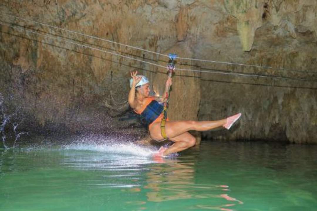 A woman ziplining over water in a cave, splashing her feet as she glides.