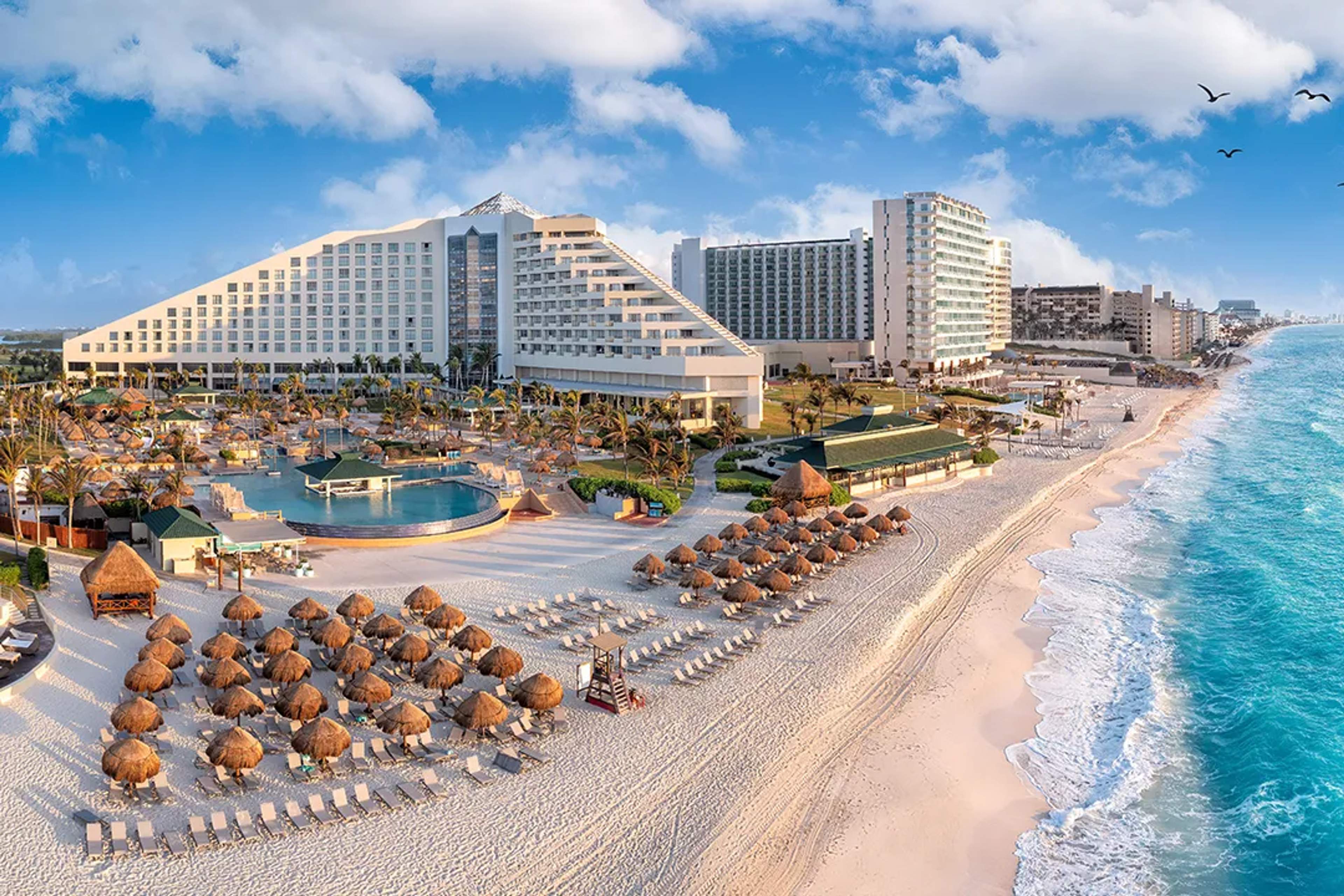Resort frente al mar en Cancún con palapas, alberca y aguas turquesa bajo un cielo despejado.