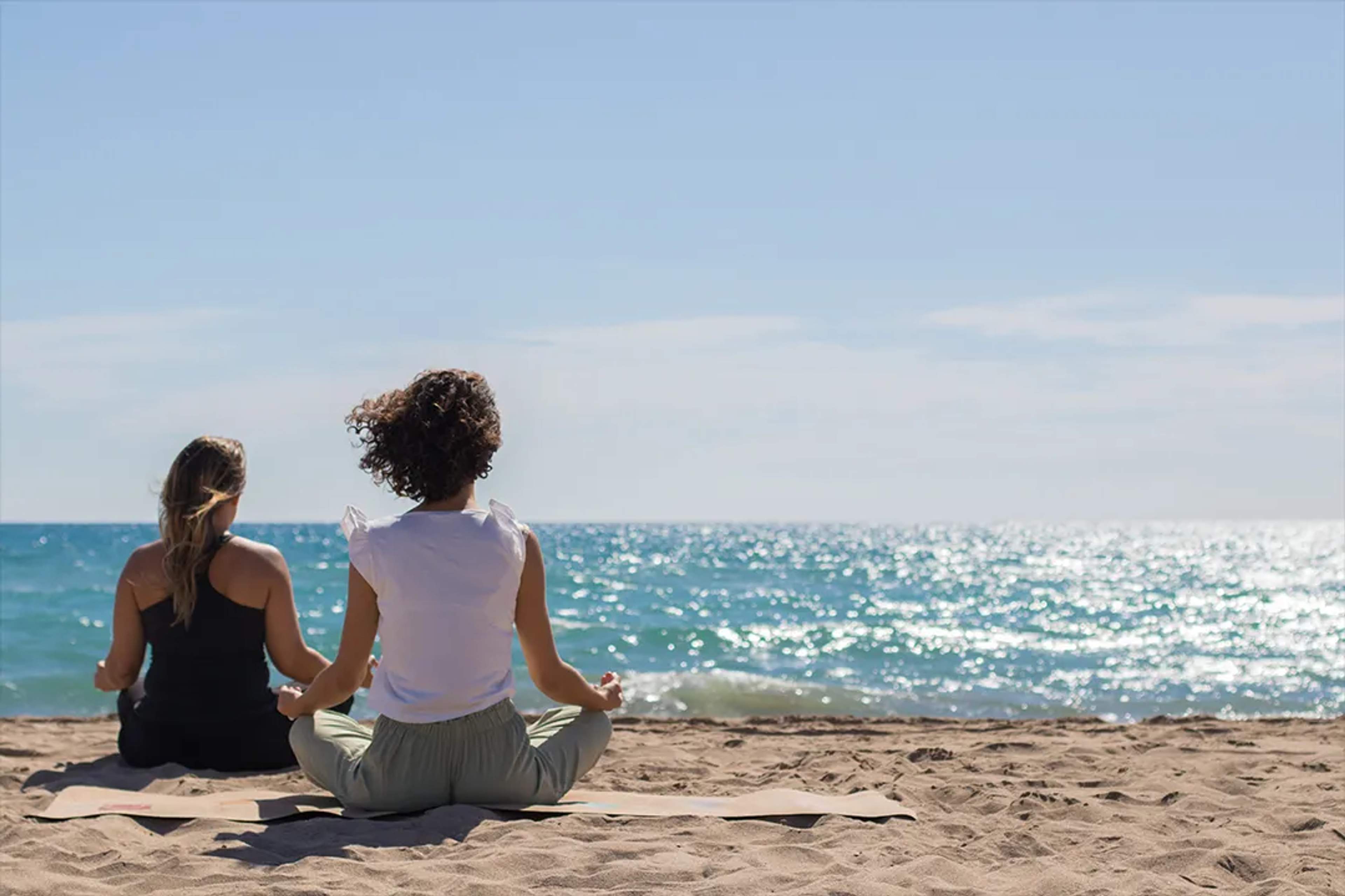 Dos mujeres practican yoga en una playa soleada con vista al mar durante una experiencia de bienestar