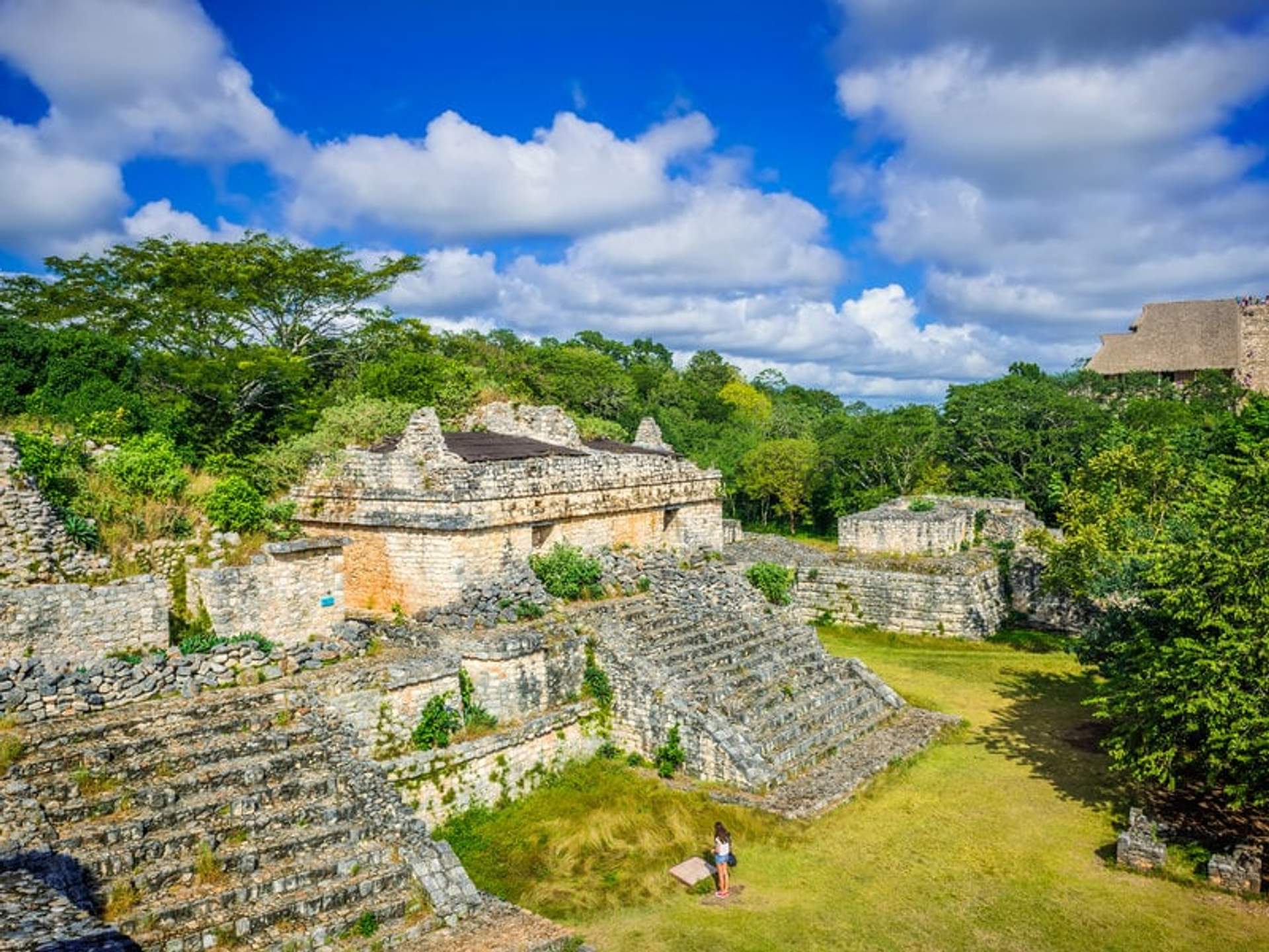 A person standing near ancient Mayan ruins at Ek Balam, surrounded by lush greenery under a bright blue sky with fluffy clouds.