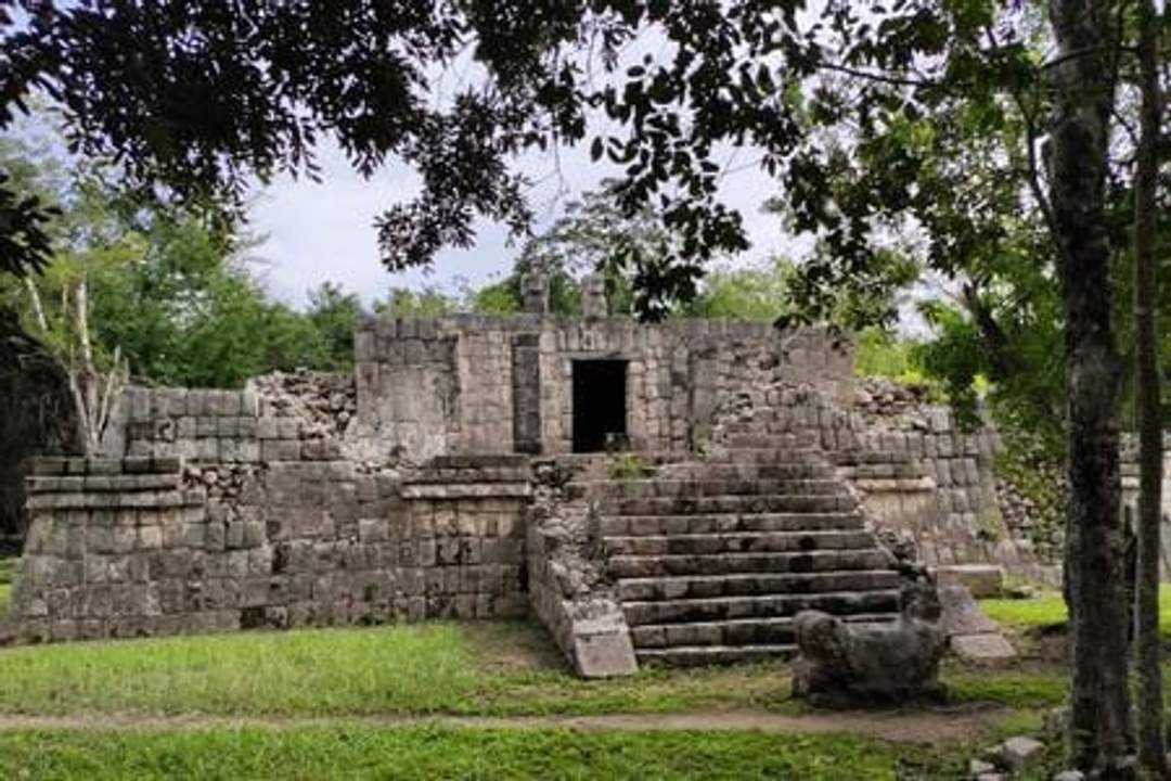 A photograph of the Temple of the Initial Series, an ancient stone structure at Chichen Itza, featuring a central staircase leading up to a platform with a doorway. The ruins are surrounded by greenery and partially shaded by trees.